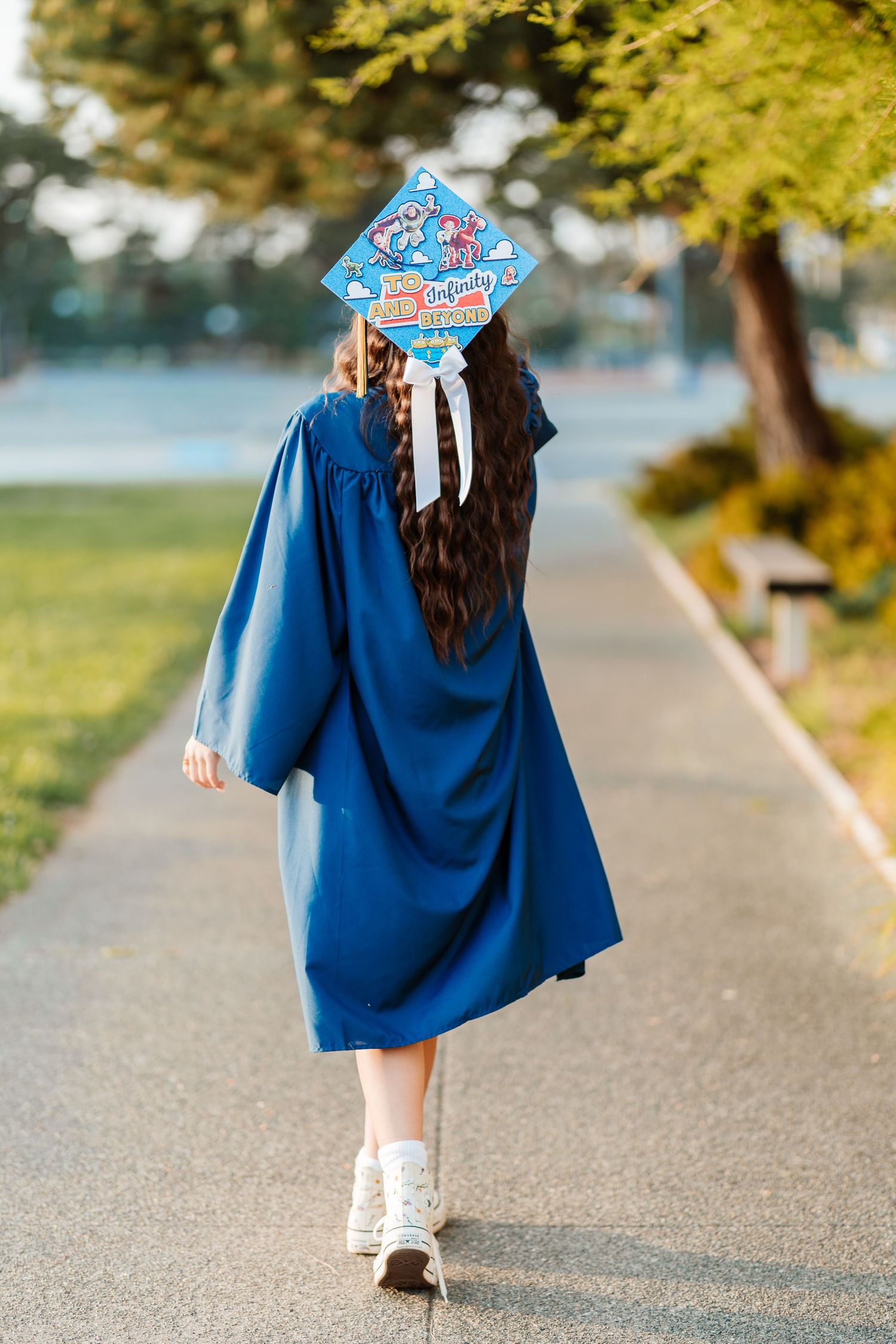 Graduate in blue cap and gown walks away down a sunny tree-lined path with decorated mortarboard.