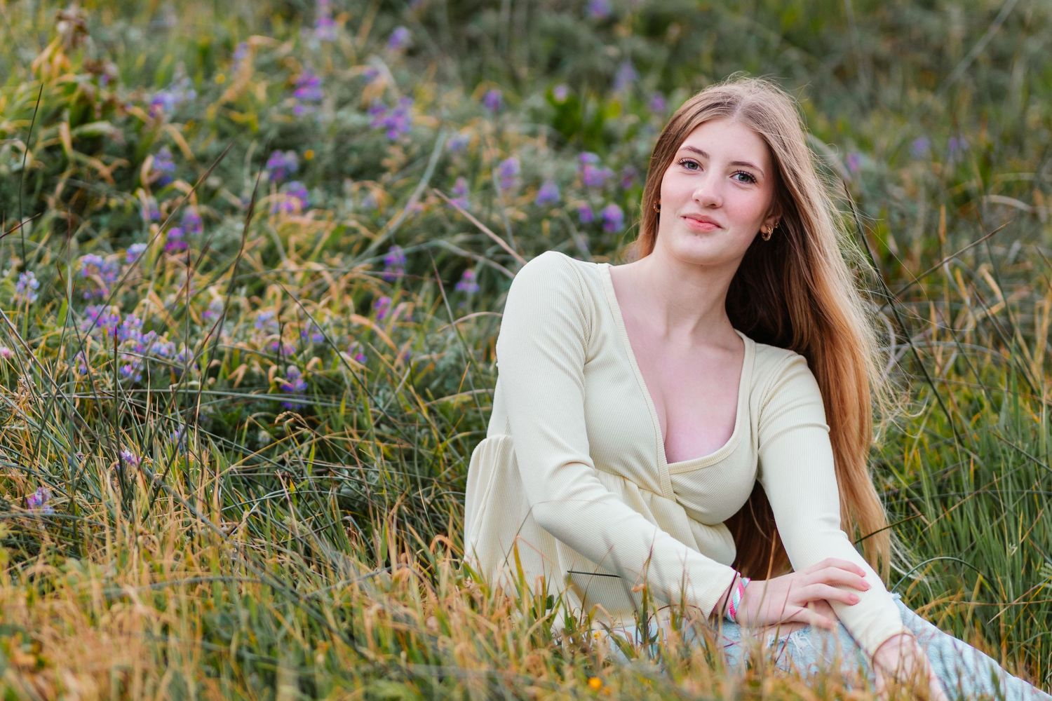 Young woman with long hair in cream shirt sitting among wildflowers in a meadow.