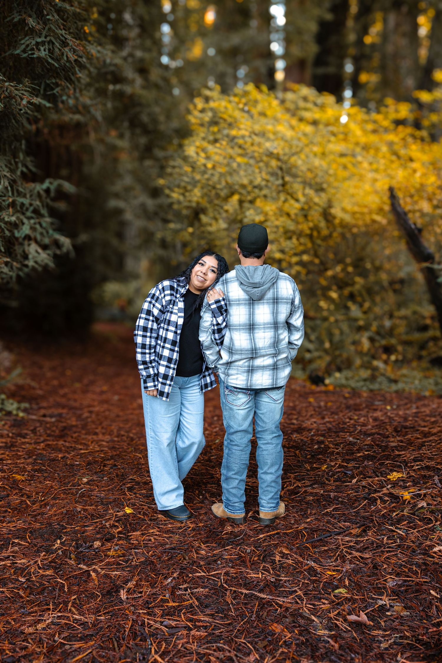 Couple in matching plaid shirts stand together on a red leaf-covered redwood path surrounded by fall foliage.