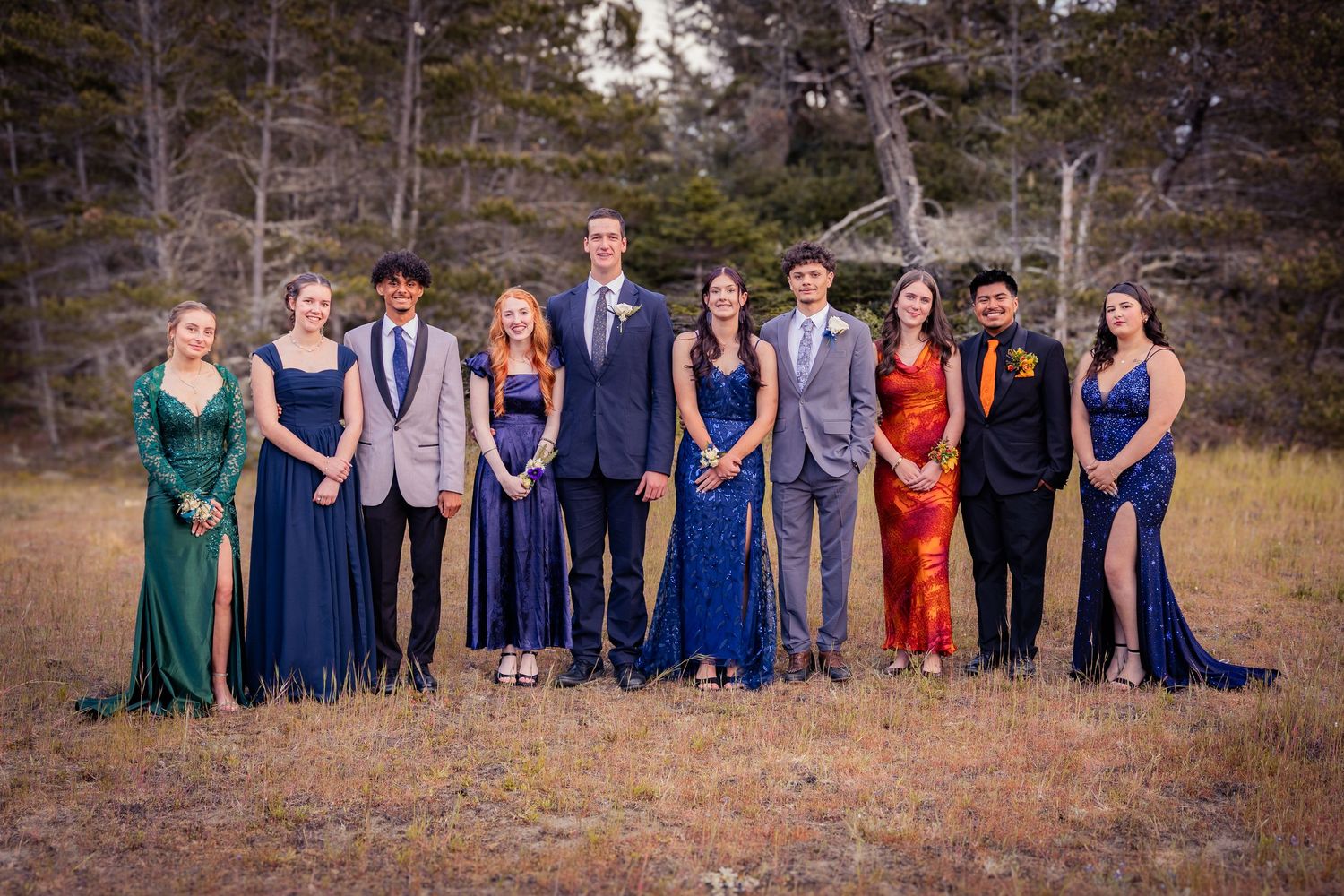 Group of ten teenagers dressed in formal prom attire posing outdoors in a field with trees in the background.
