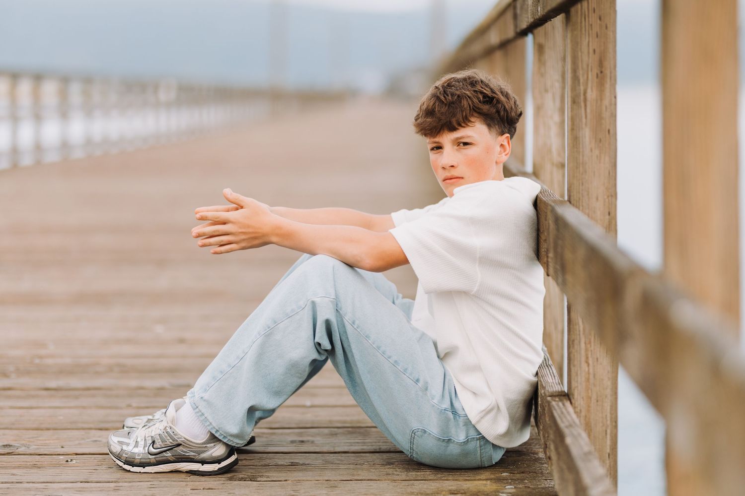 A teenage boy in white t-shirt and jeans sits relaxed on a wooden pier, arms outstretched, looking at the camera.