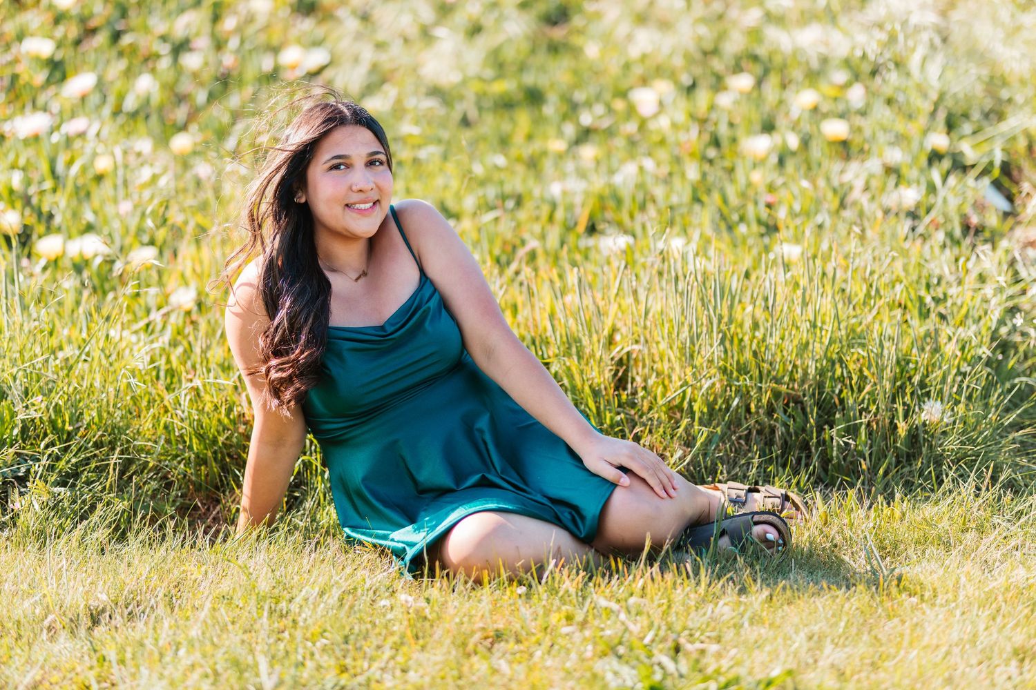 Young woman in teal dress sitting in sunny meadow with wildflowers, smiling and holding a vintage camera.