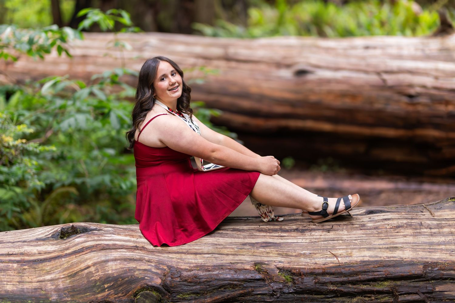Young woman in red dress sitting on fallen log in lush forest setting, smiling at camera.