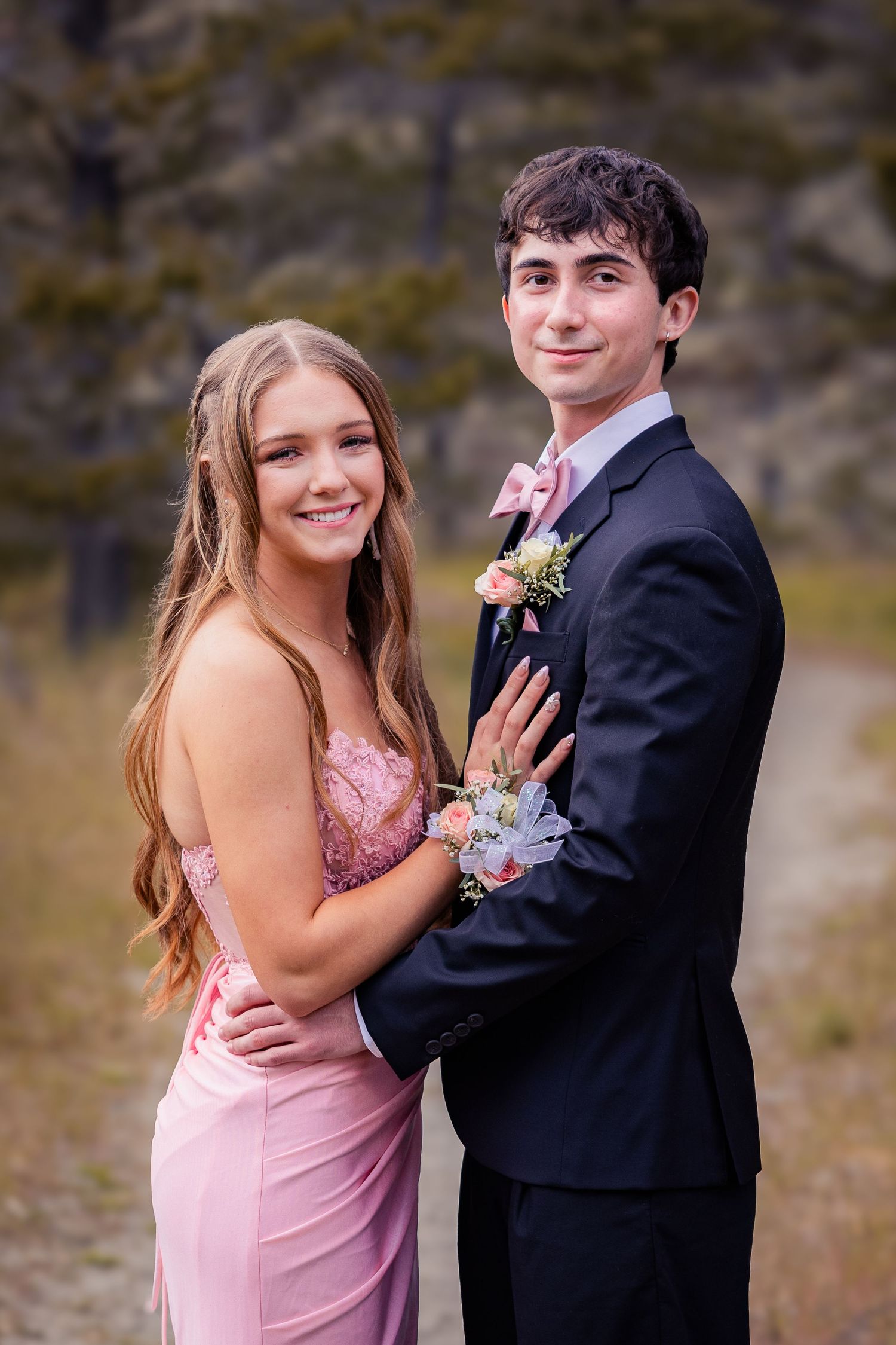 A young couple dressed in prom attire, girl in pink gown with corsage, boy in dark suit with boutonniere, posing outdoors.