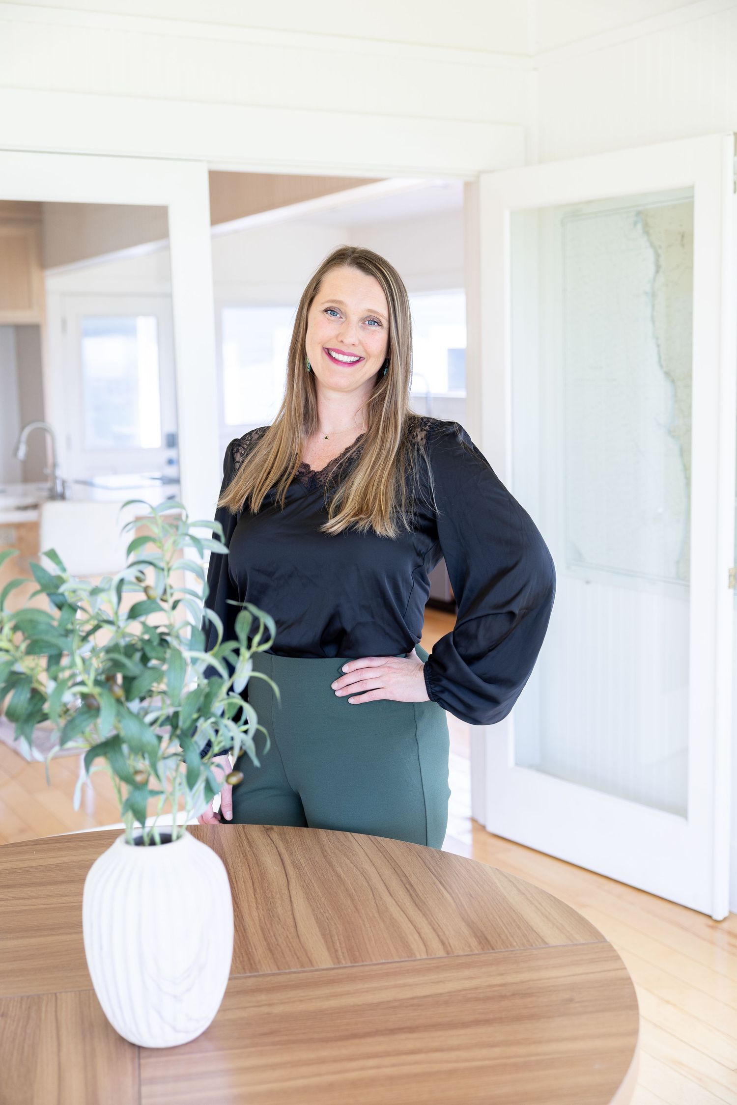 Woman in black blouse and green pants smiling confidently in a modern bright home interior with wooden table.
