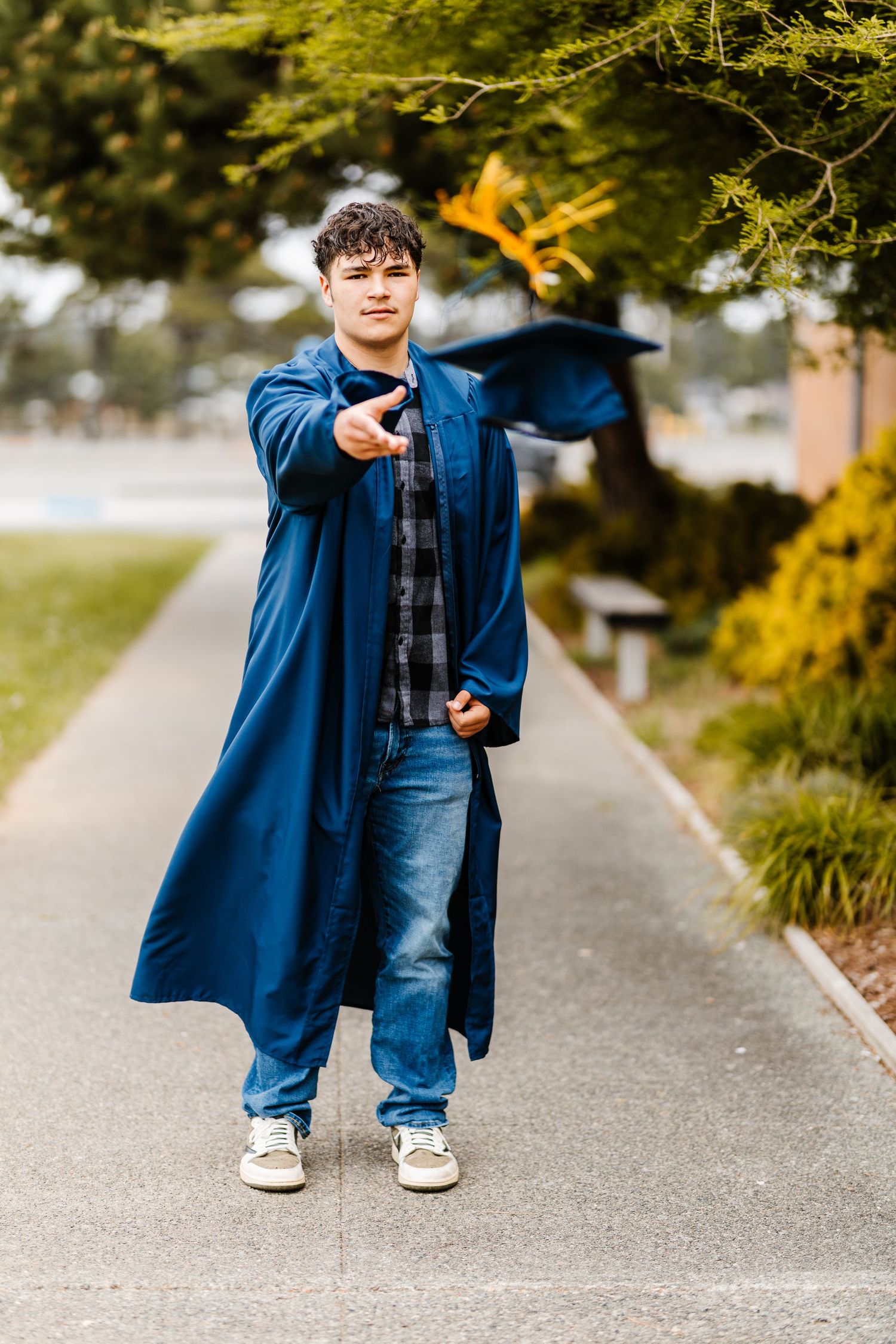 Young man in blue graduation gown pointing at camera while tossing cap outdoors on a tree-lined path.