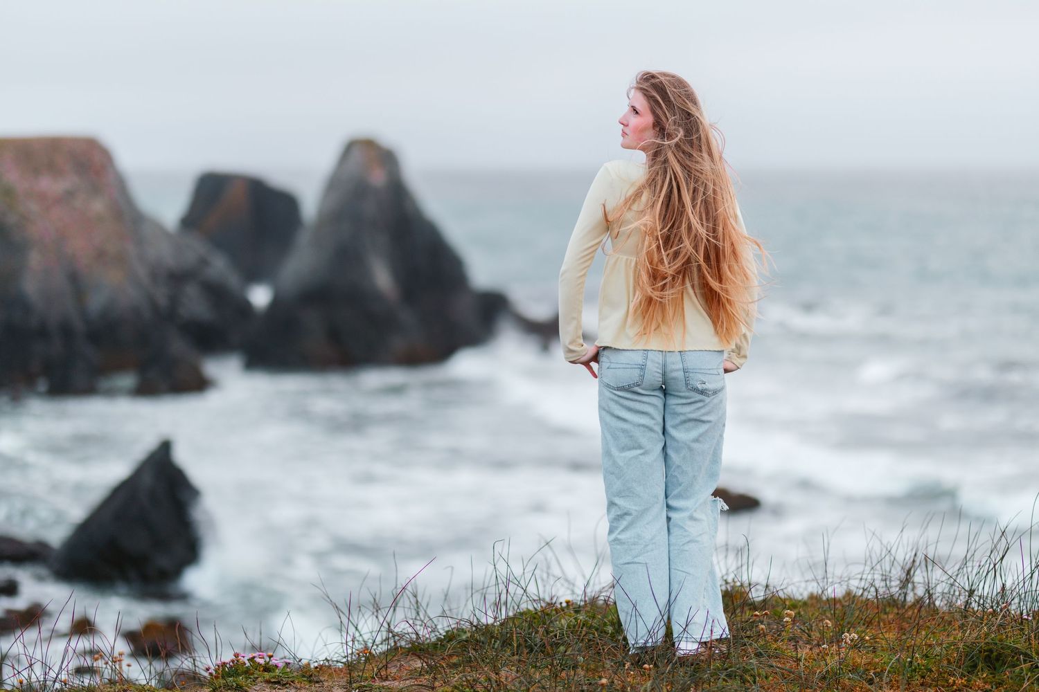 Young woman with long blonde hair standing on coastal cliff overlooking rocky ocean shore.