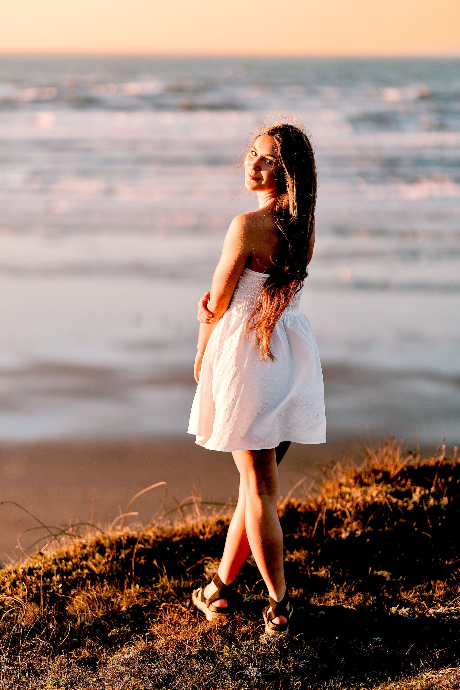 Young woman in white strapless dress stands on coastal bluff at sunset with ocean waves behind her.