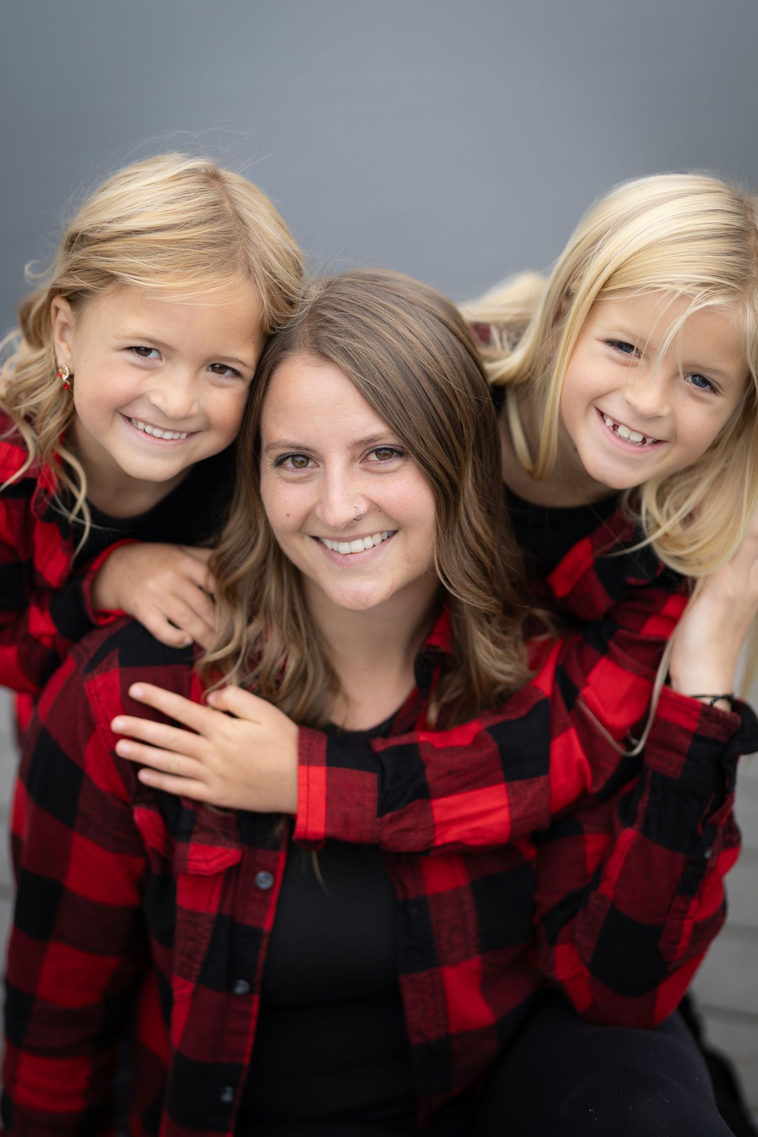 Smiling mother with two blonde daughters in matching red plaid shirts against gray background.