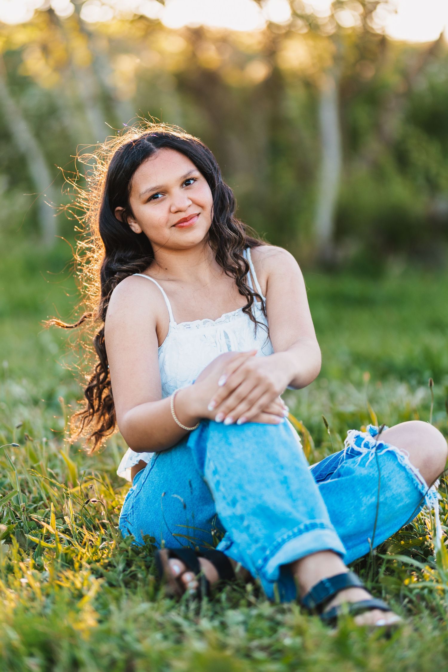 Young woman with long dark curly hair sits cross-legged on green grass wearing white top and jeans in golden hour light.