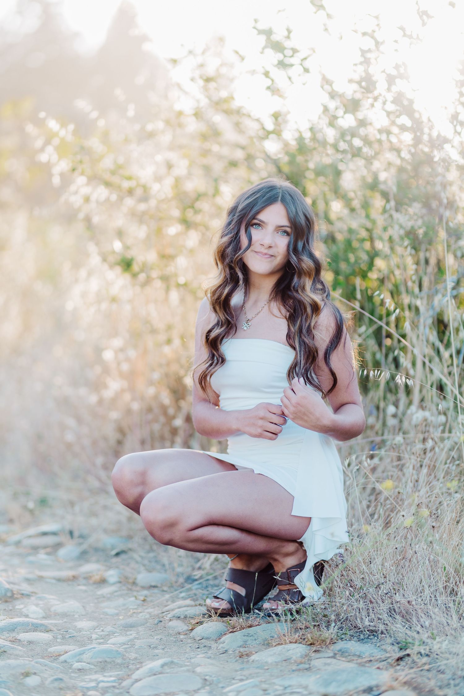 Young woman with long wavy hair in white dress crouching on rocky path surrounded by golden wild grass in sunlight.