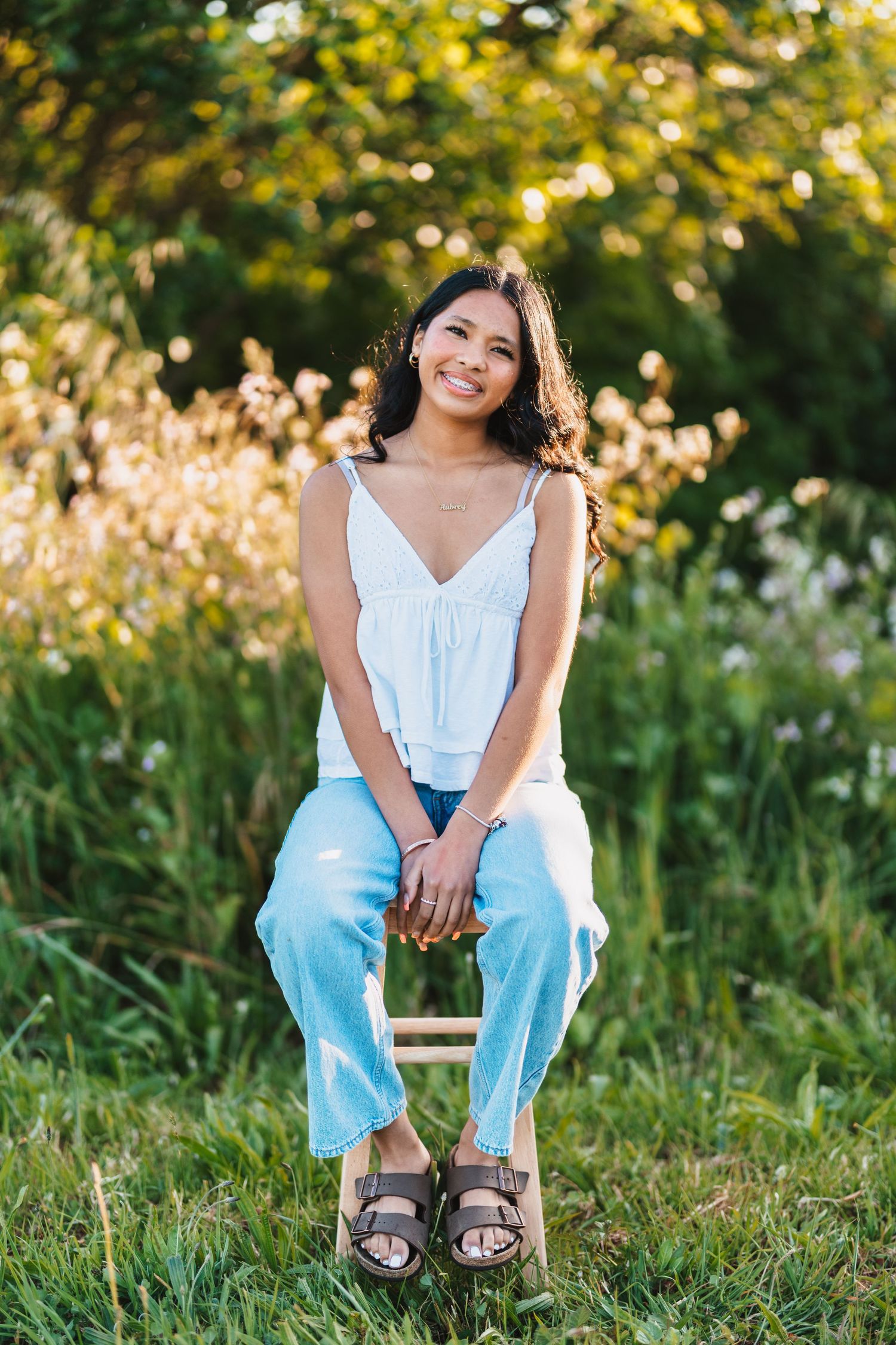 Young woman smiling outdoors, sitting on stool in white tank top and blue jeans with sandals, green nature background.
