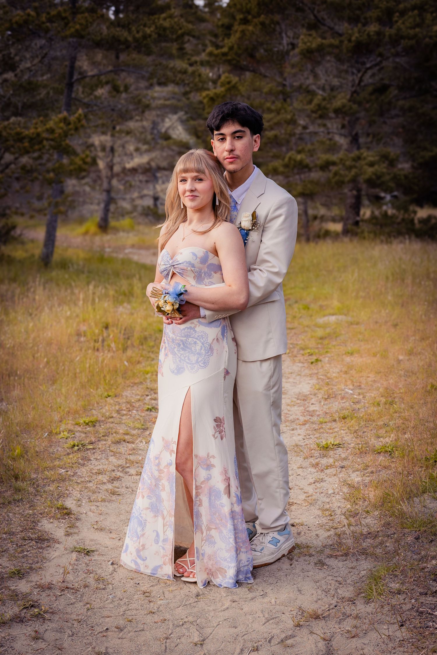 Young couple in prom attire pose outdoors, girl in floral gown holding bouquet, boy in tan suit embracing her.