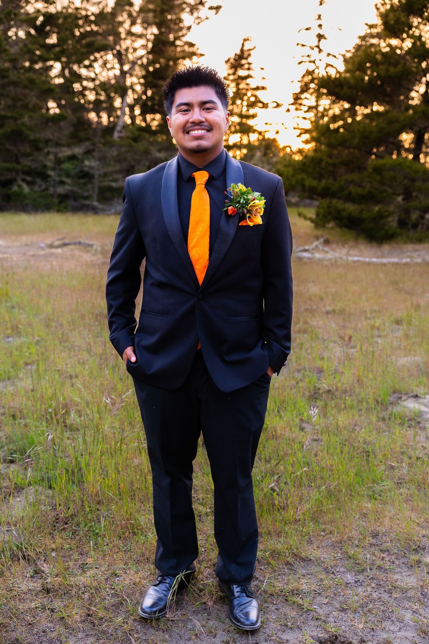Young man in a black tuxedo with orange tie and boutonniere smiles at sunset in a grassy field.