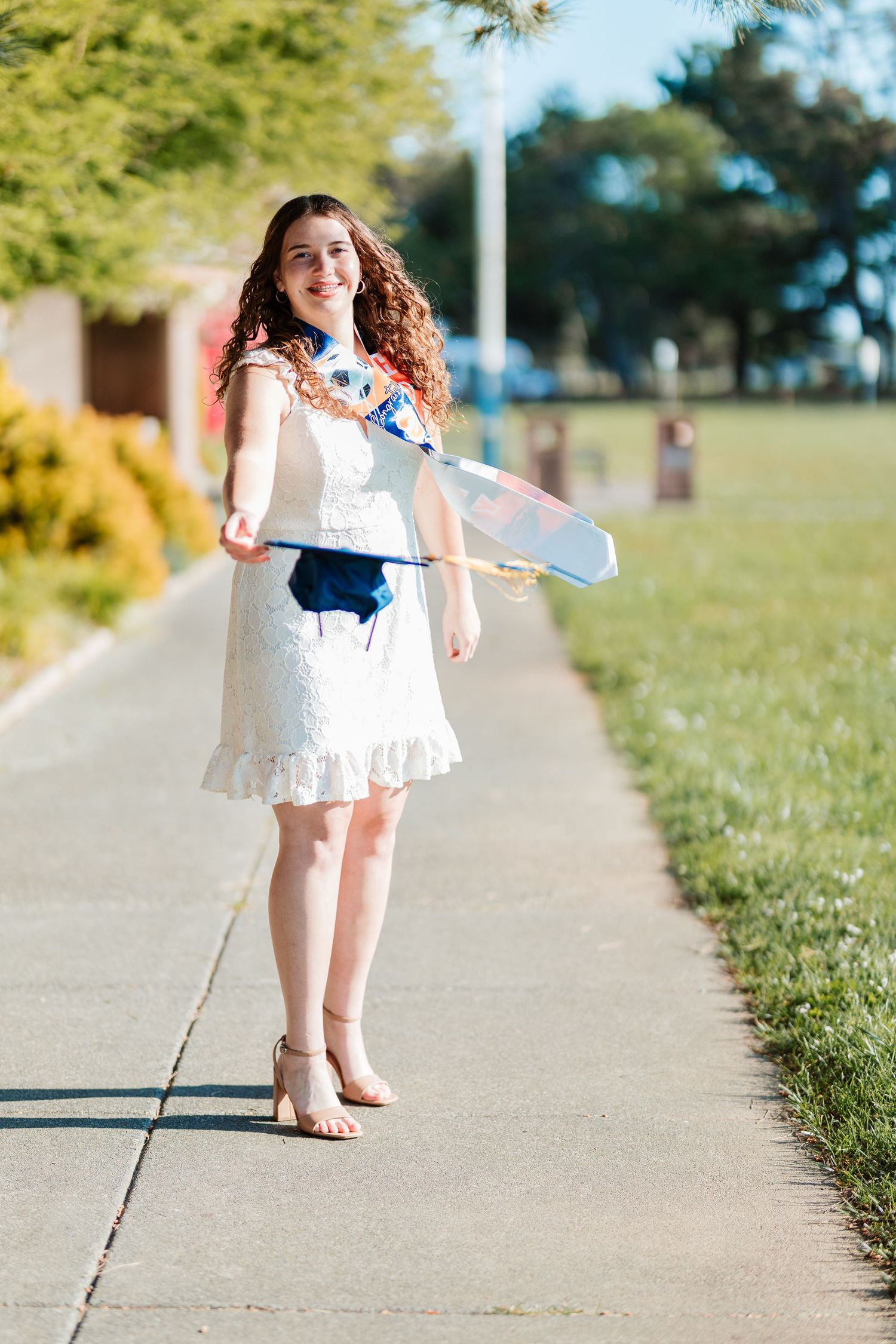 Young woman in white dress holding graduation cap on sunny sidewalk, celebrating academic achievement outdoors.