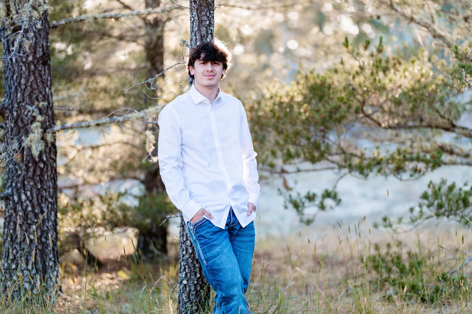 A Senior high school boy in a white shirt and jeans leans casually against a tree outdoors.