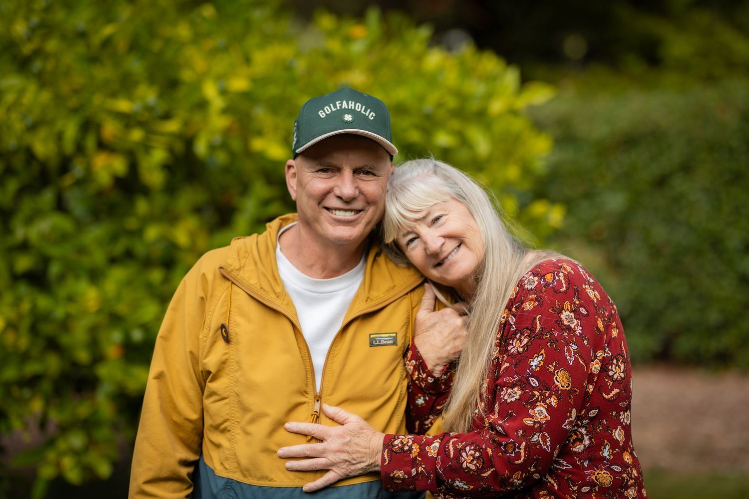 Happy mature couple posing together outdoors, man wearing yellow jacket and green cap, woman in red floral blouse.