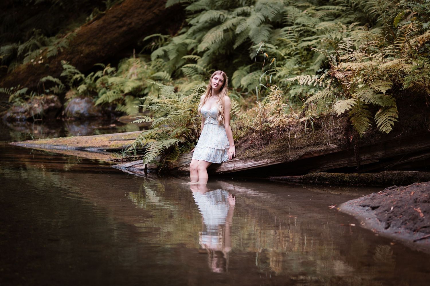 Young woman in a white dress standing in a reflective forest stream surrounded by ferns.
