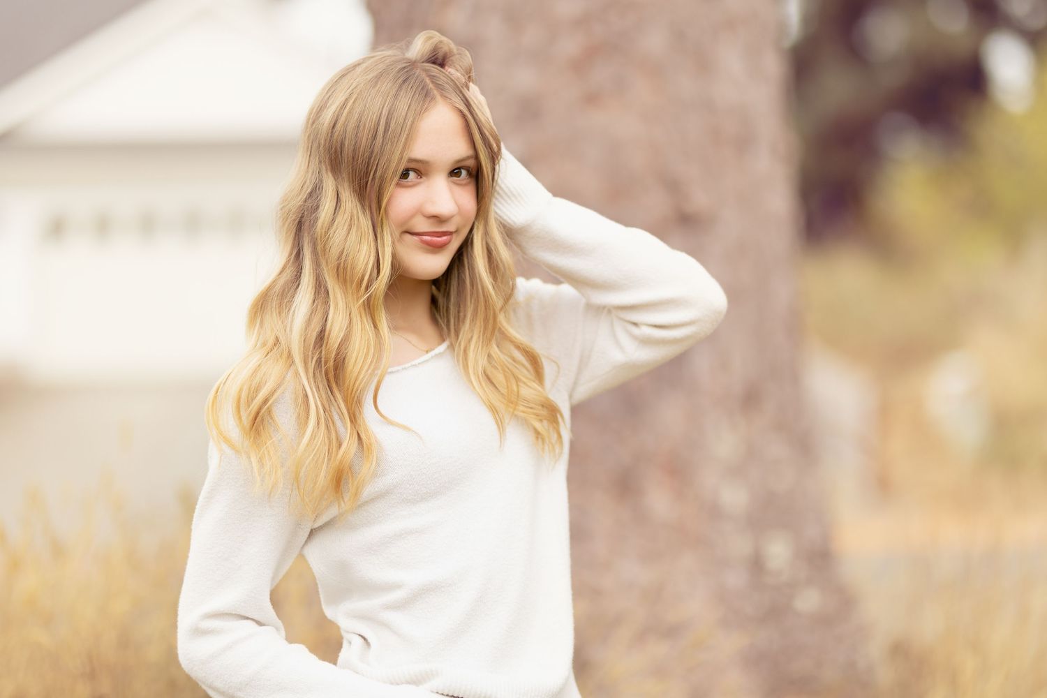 Blonde young woman in white sweater posing outdoors with hand in hair.