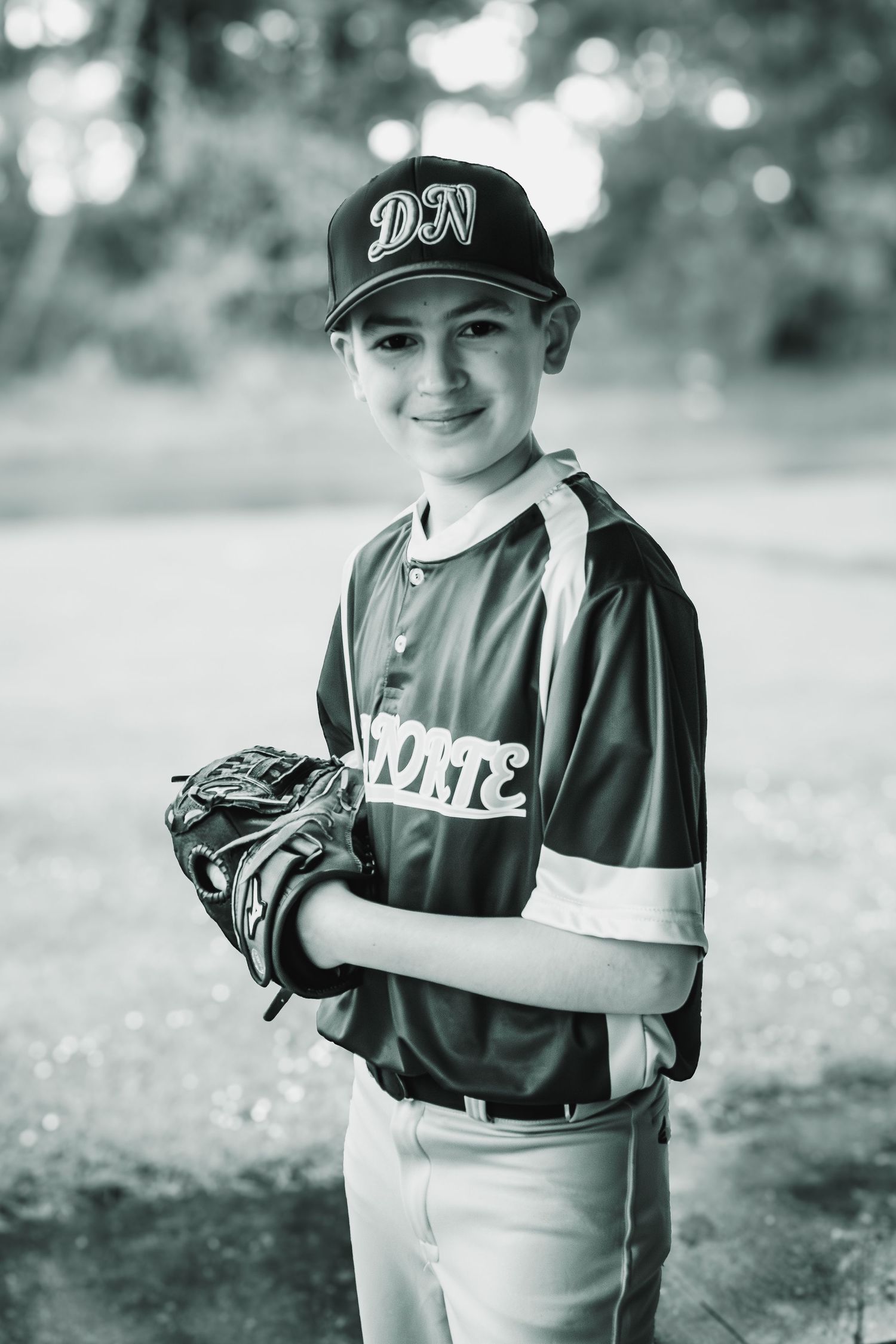 Black and white portrait of smiling boy in DN baseball cap and Norte jersey holding glove.