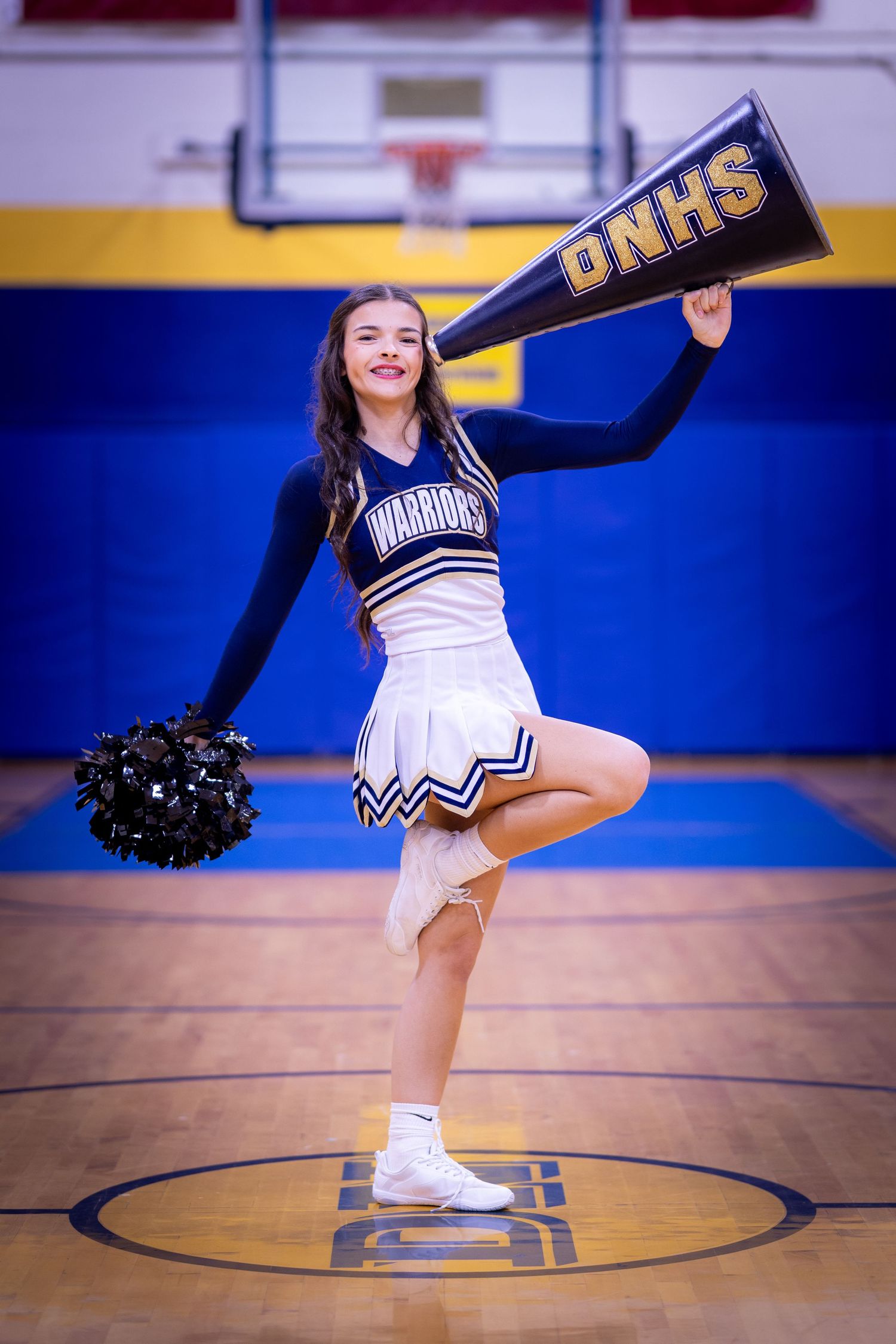 Cheerleader in Warriors uniform holding DNHS pennant and pom-pom on a gymnasium court.