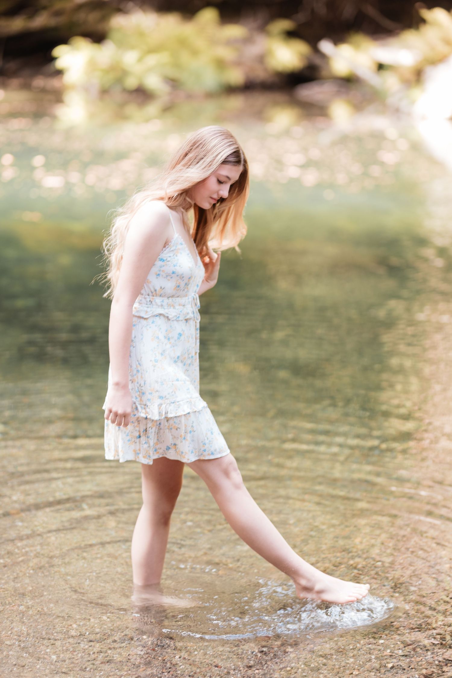Young woman in white floral dress wading barefoot in a shallow sunlit creek.