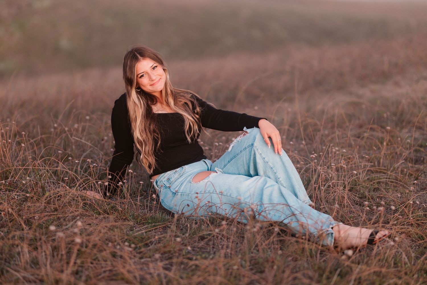 Teen girl in black top and blue floral jeans sitting relaxed in a golden dry grass field.