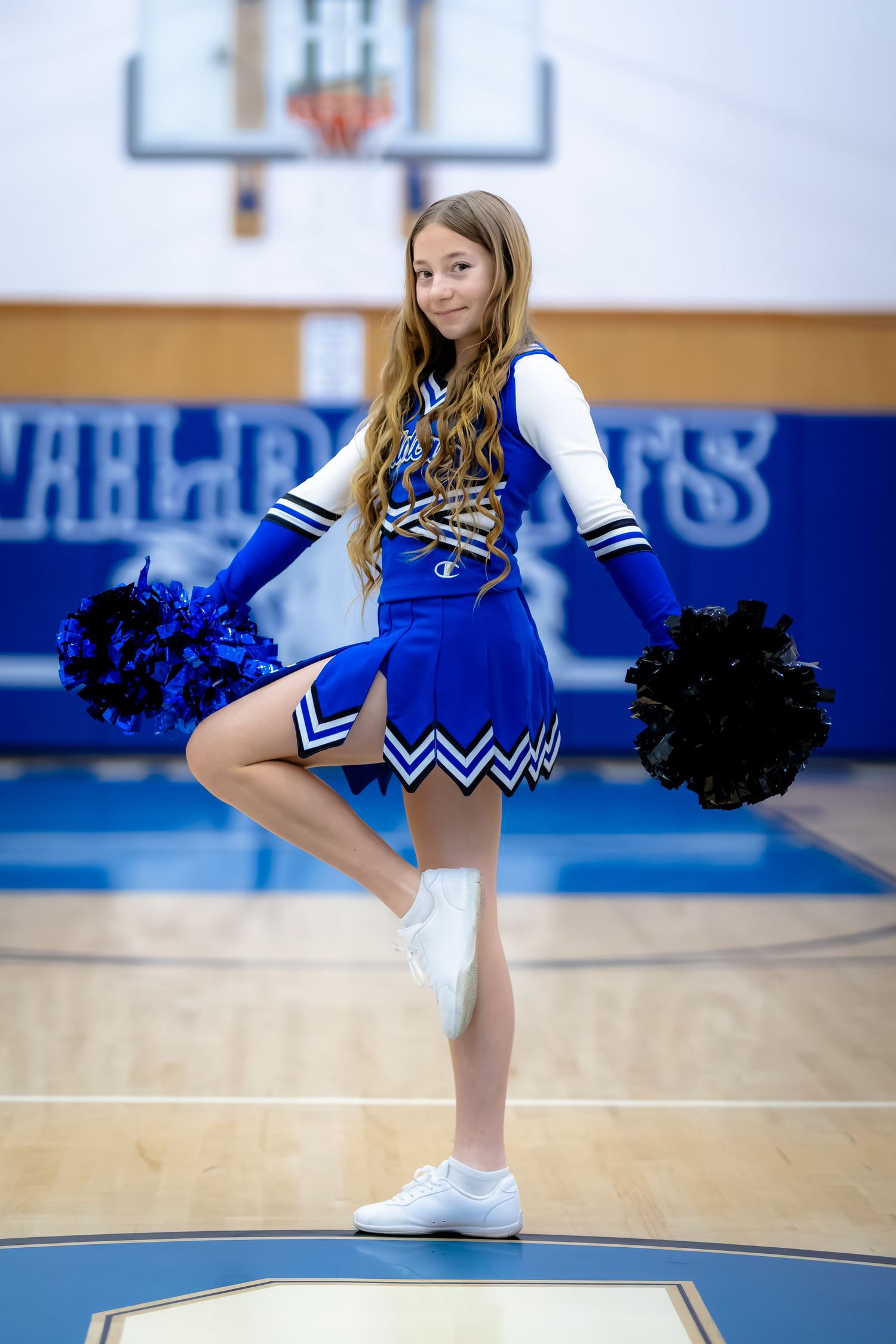 Young cheerleader in blue Wildcats uniform posing with pom-poms raised leg in school gymnasium.