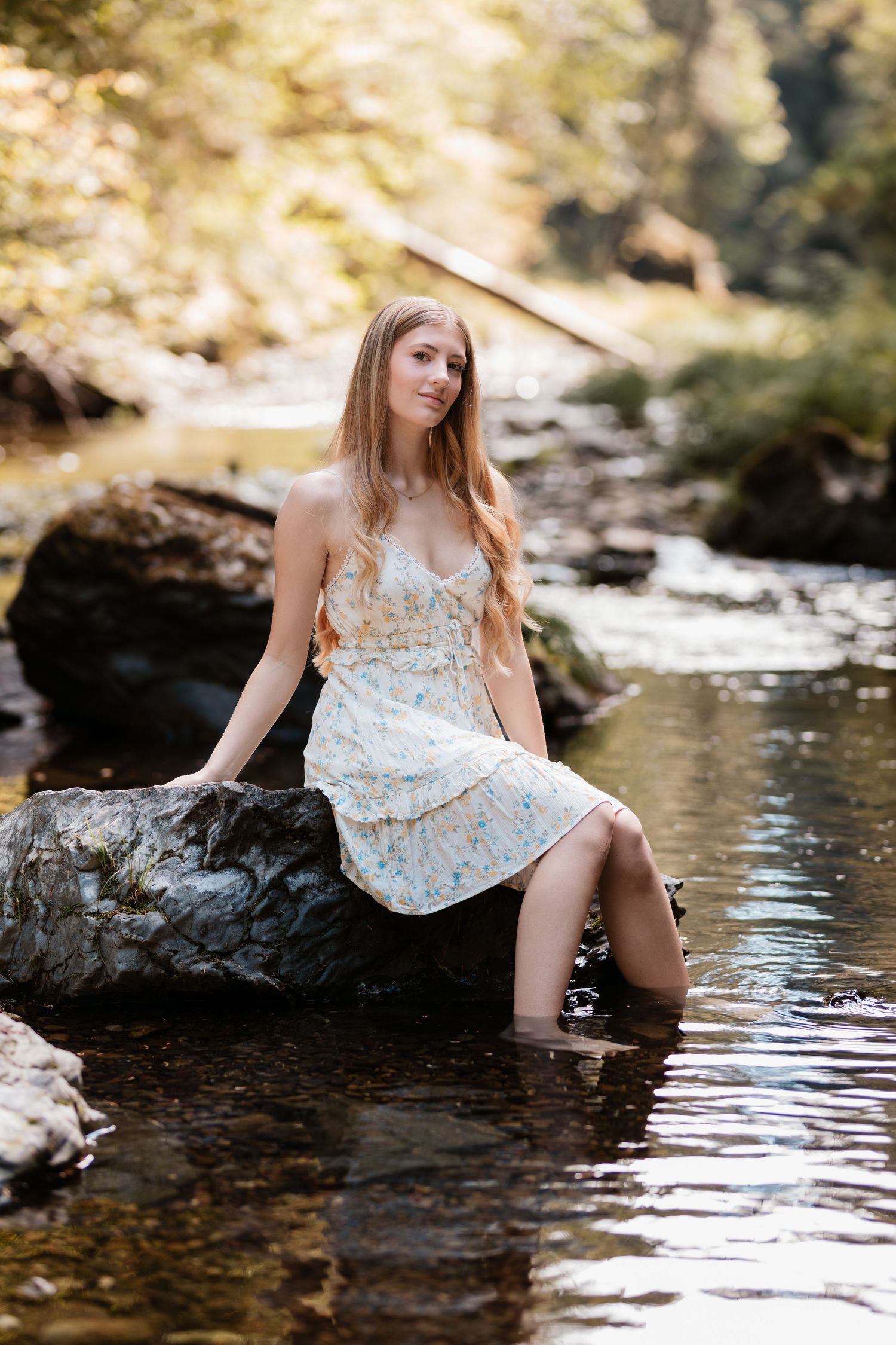 Young woman in a floral sundress sitting on a rock in a shallow sunlit forest stream.