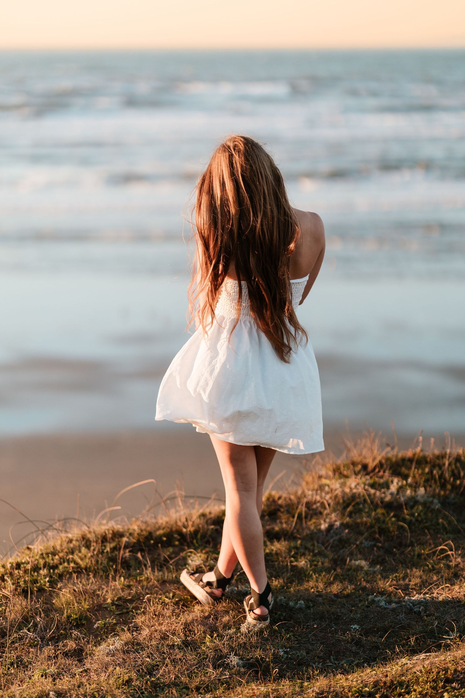 Young woman in a white dress standing on a grassy cliff overlooking ocean waves at sunset.