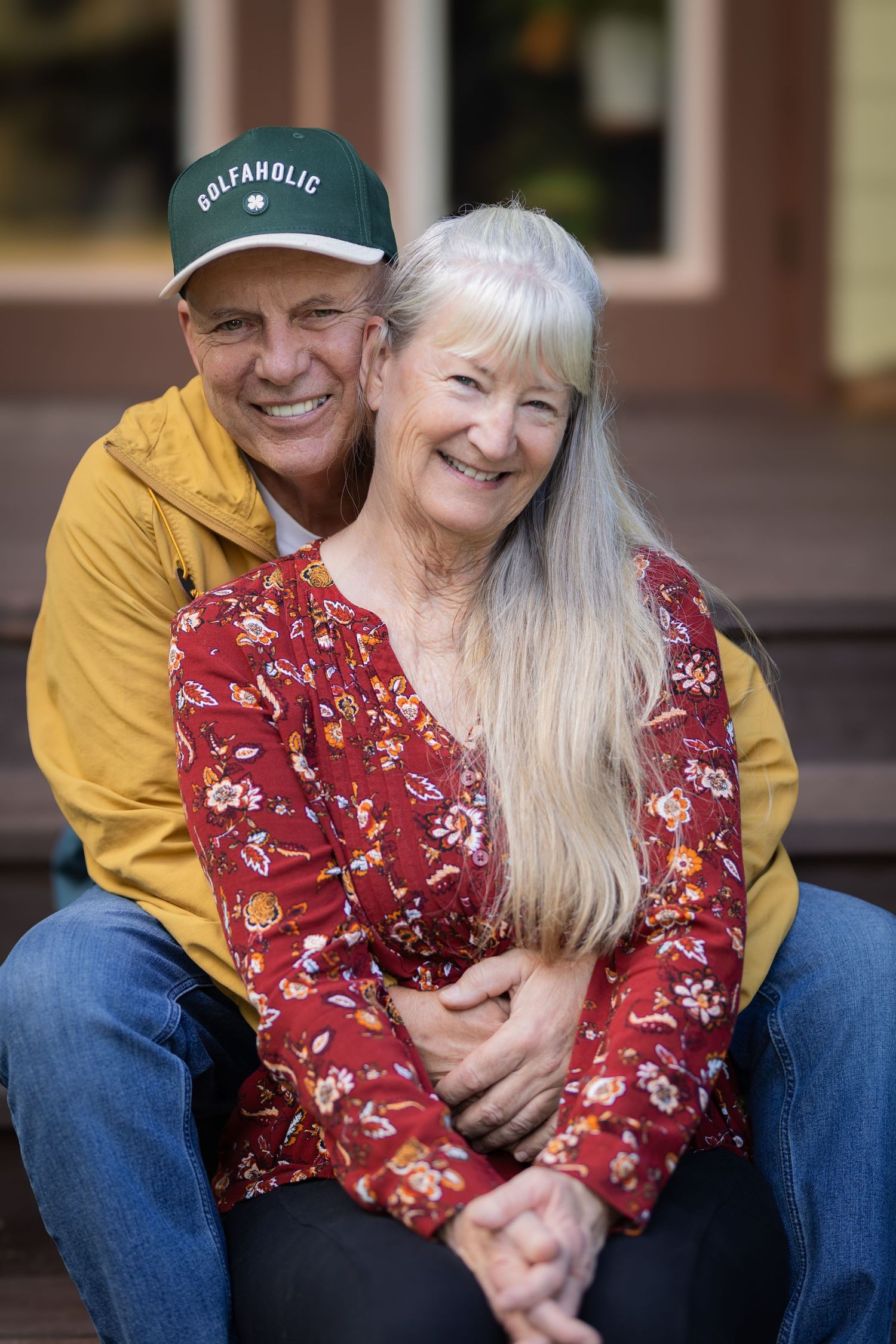 Joyful senior couple smiling closely together, man in yellow jacket and Golfaholic cap hugging woman in red top.