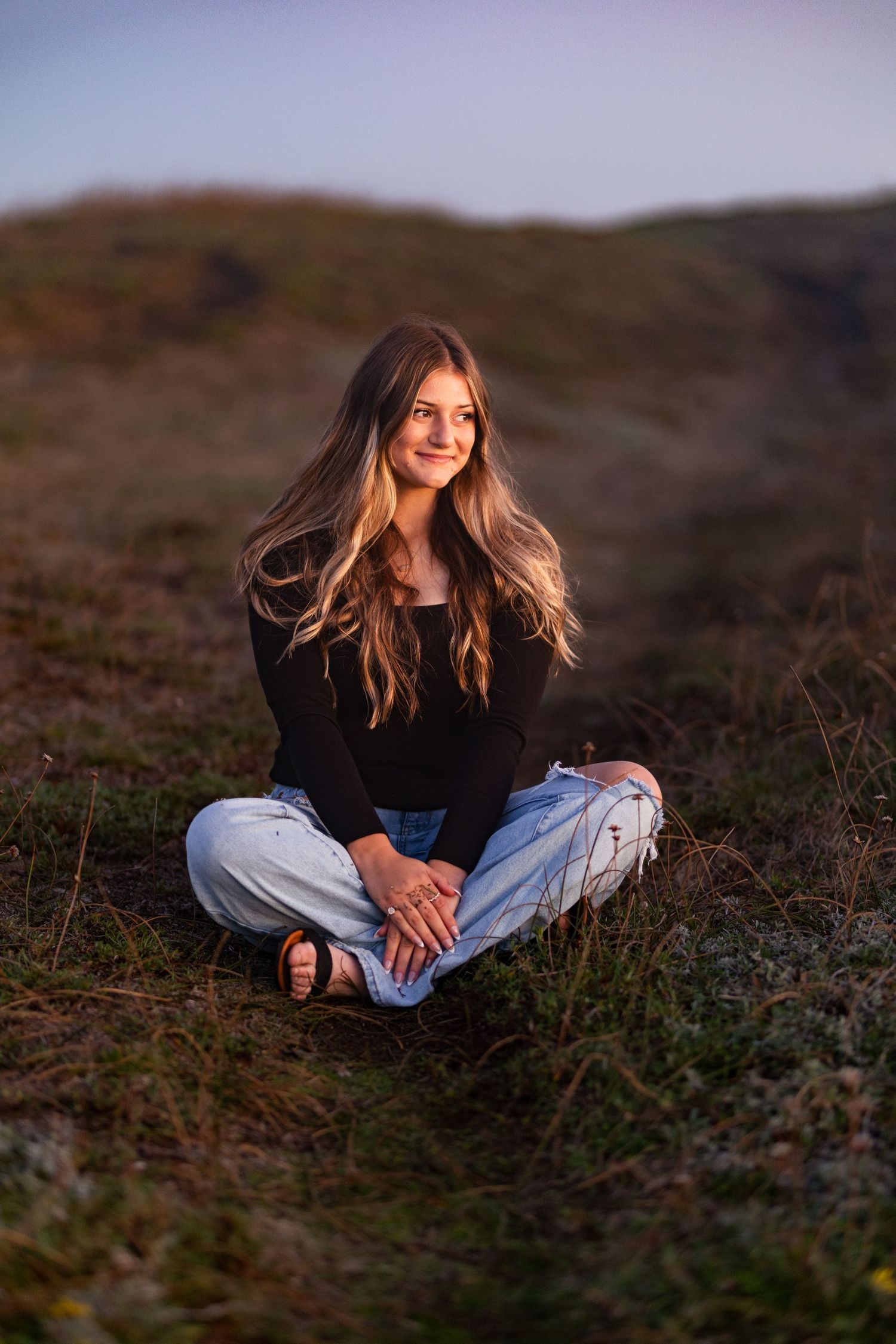 Teen girl in black top and ripped jeans sitting cross-legged on grassy hillside at sunset.