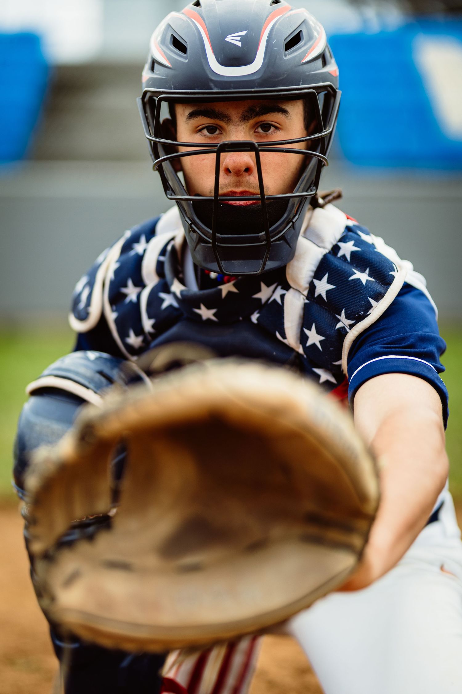 Close-up of baseball catcher in American flag chest protector and helmet extending mitt forward.