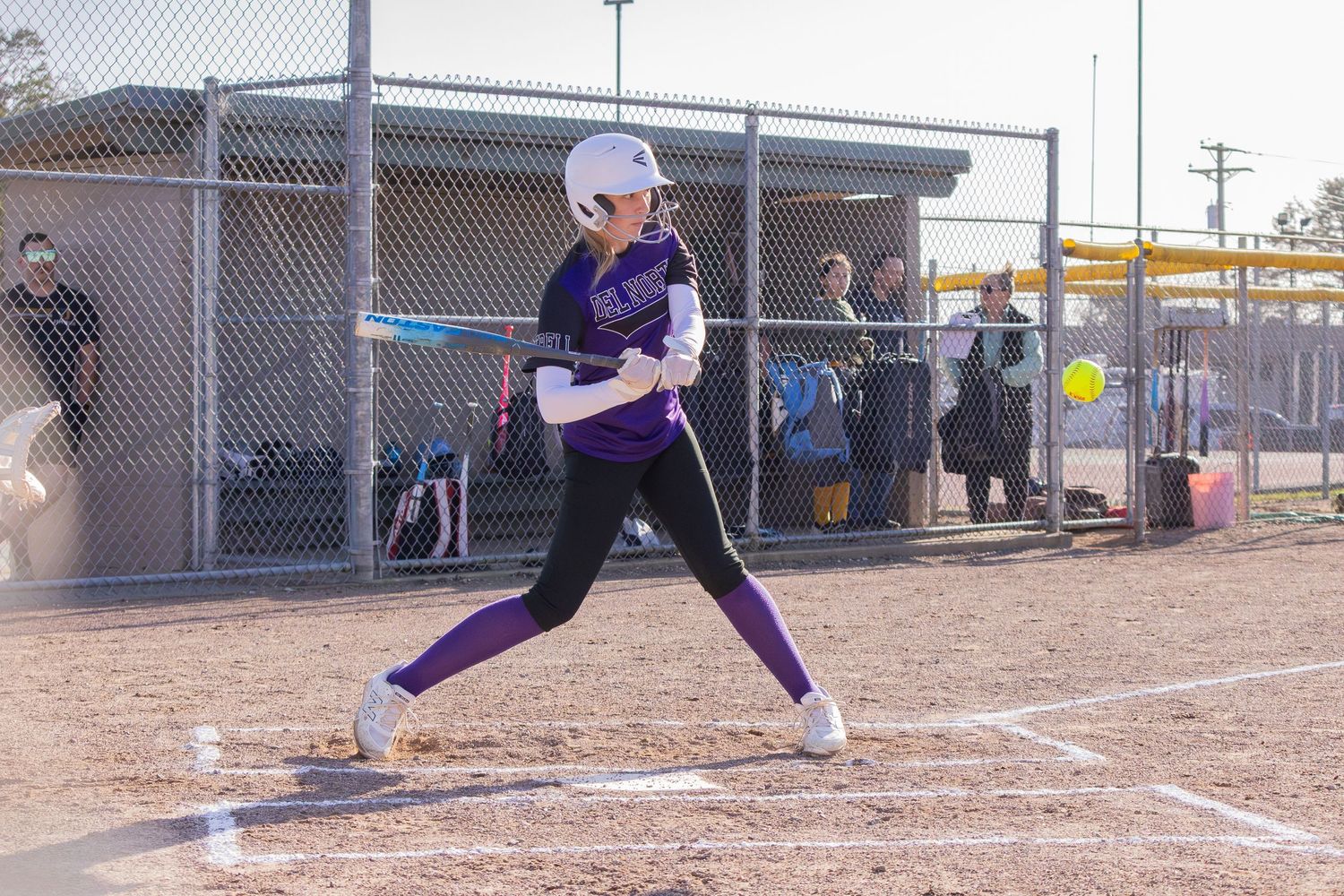 Female softball player in purple uniform swings bat during a competitive game on a dirt baseball diamond.