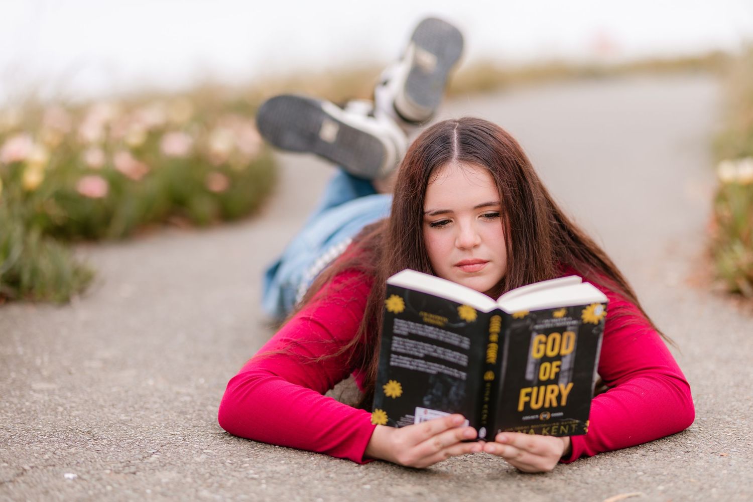 Teen girl in pink top lying on pavement reading God of Fury book during an outdoor 8th grade photo session.