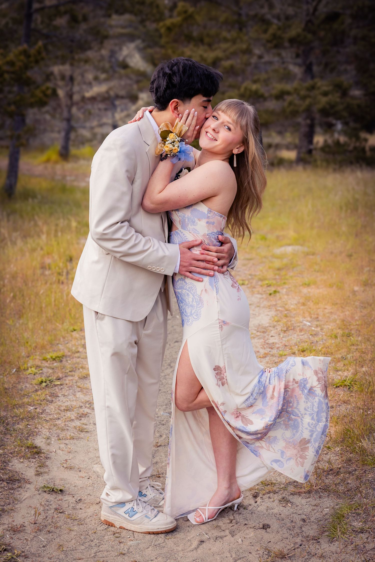 Young couple in formal prom attire embrace outdoors, boy in tan suit kisses girl in floral dress.