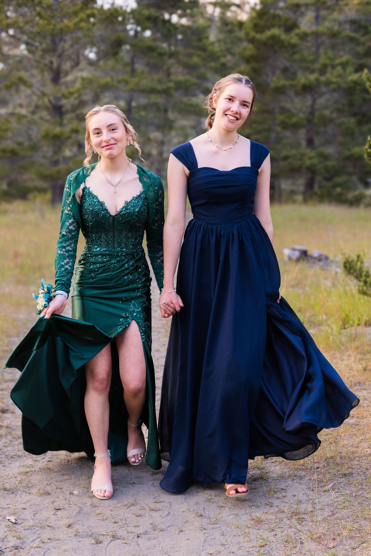 Two smiling young women in green sequin and navy blue prom dresses walk hand in hand outdoors.