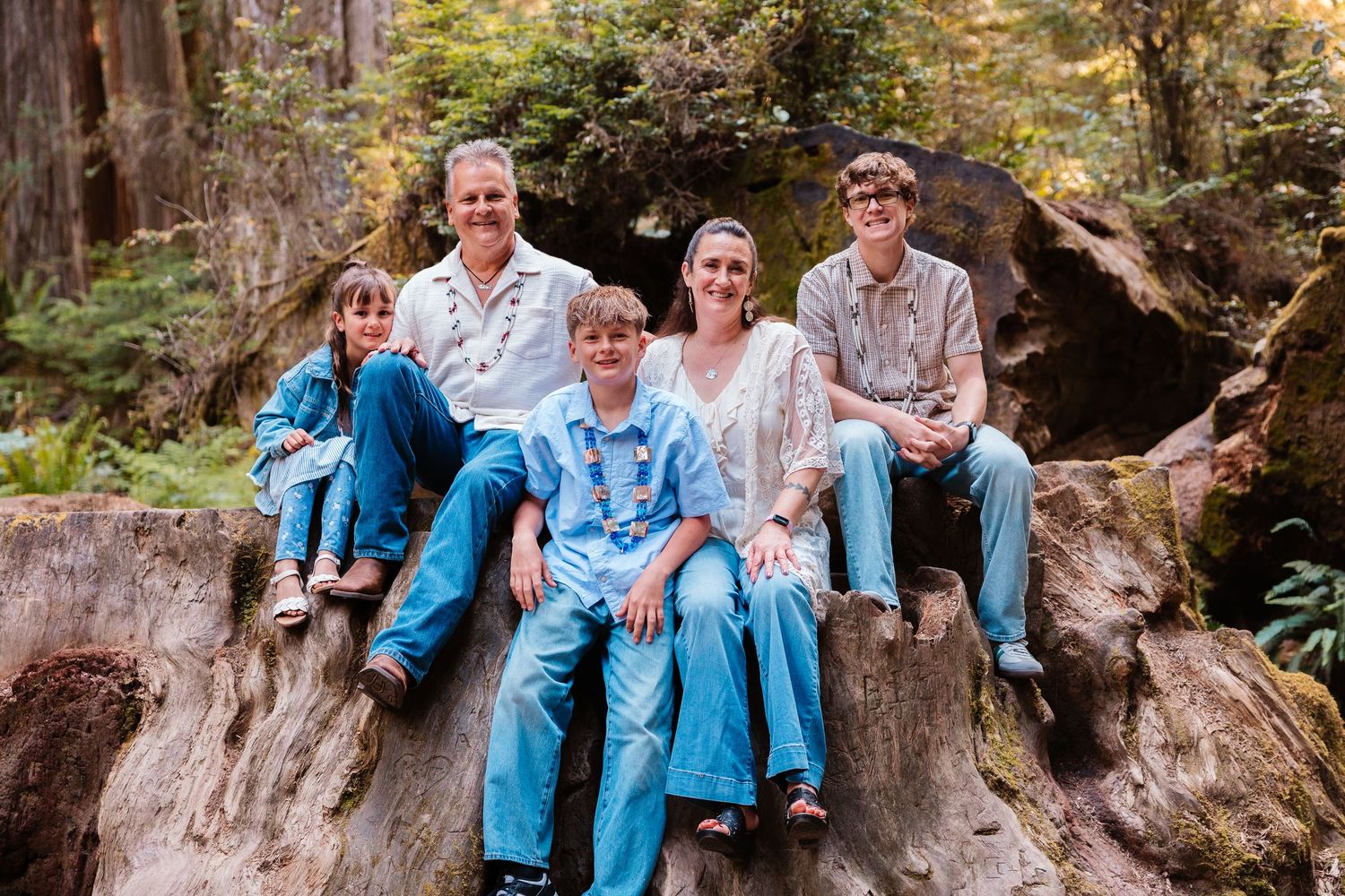 Family of five smiling together on a large tree stump in a lush forest setting.