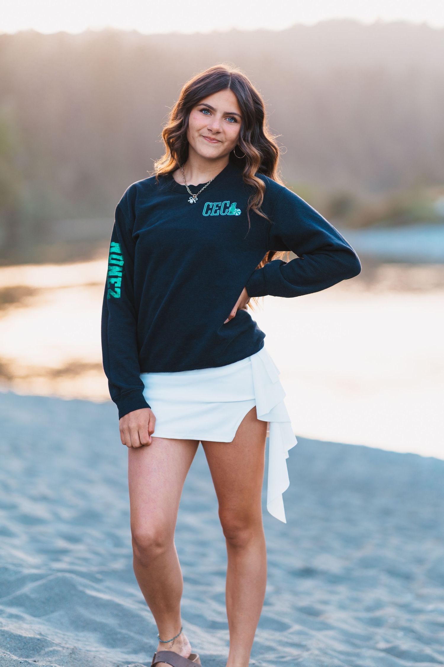 Young woman in black long-sleeve shirt and white skirt posing on sandy beach at golden hour.