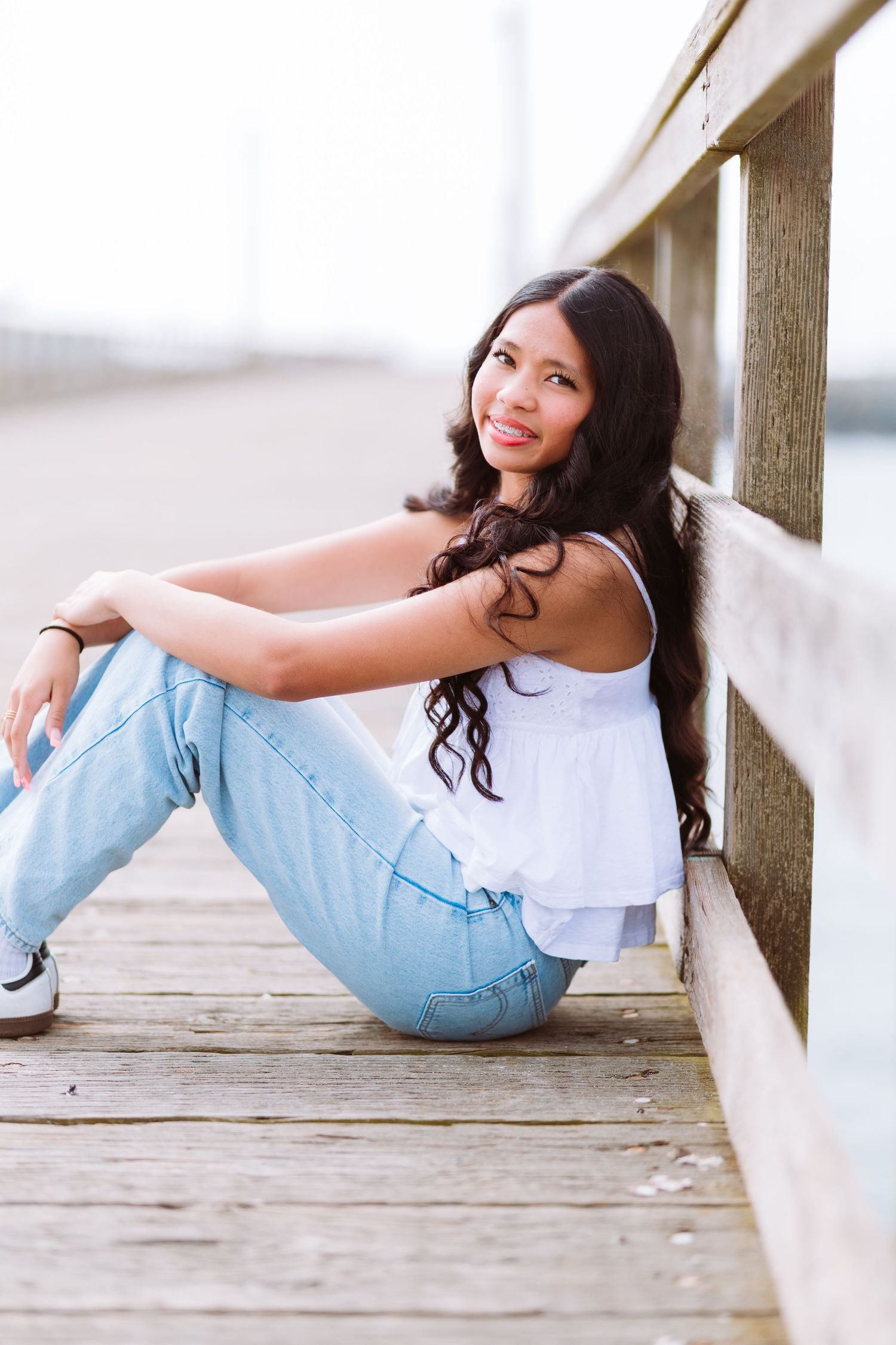 Young woman with long curly hair sits on a wooden pier, wearing a white top and light blue jeans, smiling.