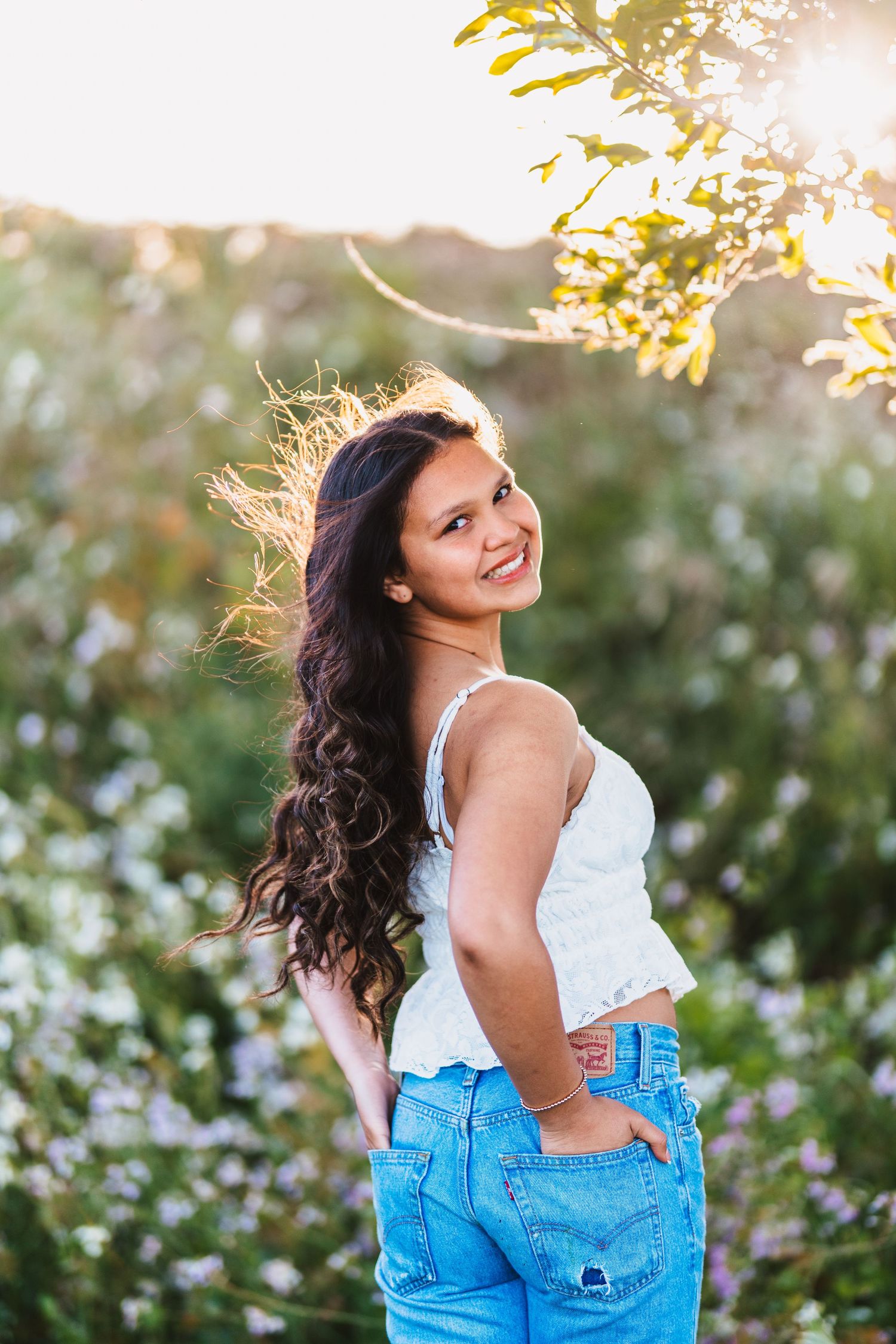 Young woman with long dark hair smiling outdoors in white top and blue jeans among wildflowers at golden hour.