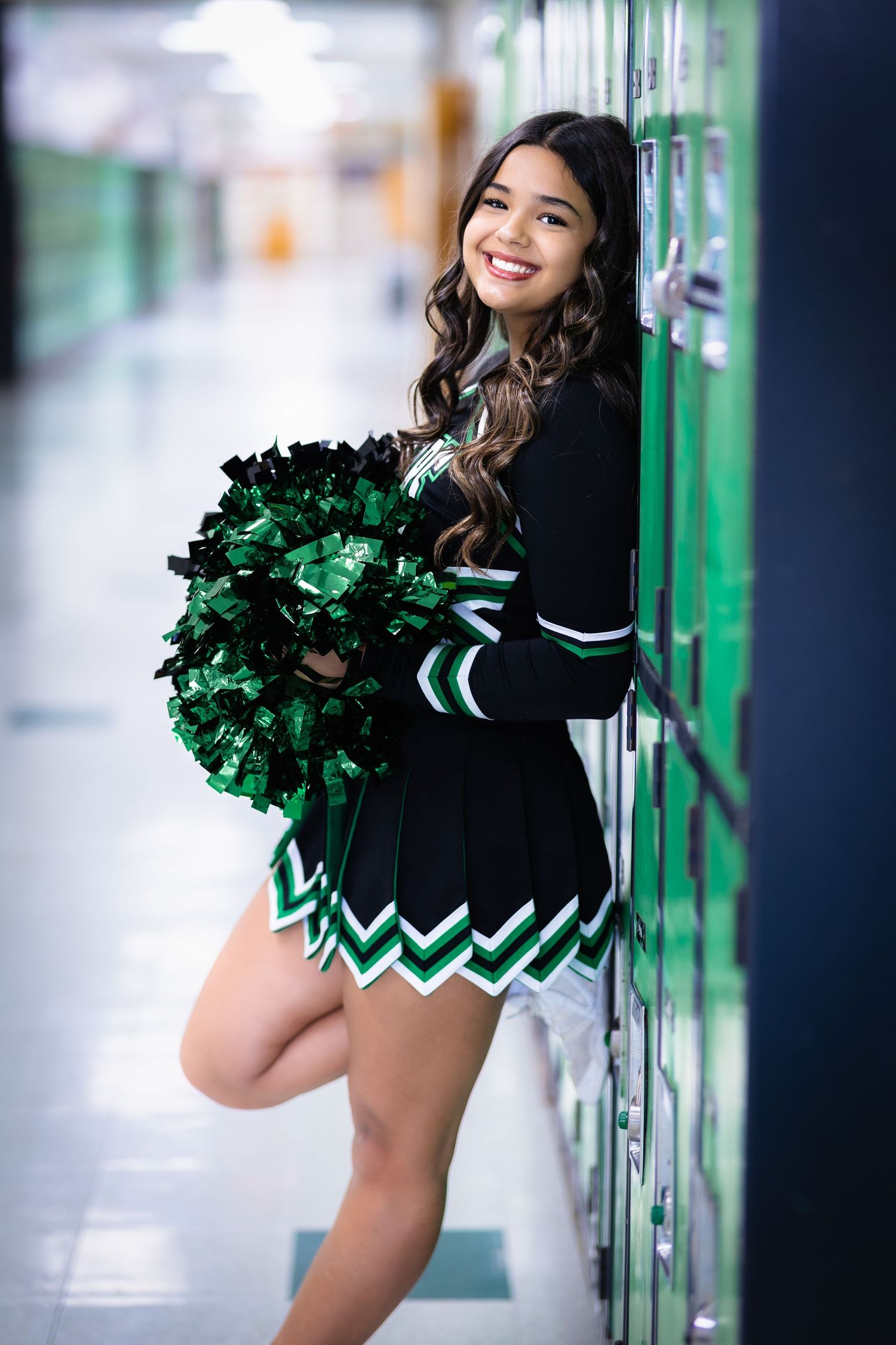 Cheerleader in black and green uniform posing with pom-poms against green lockers in school hallway.