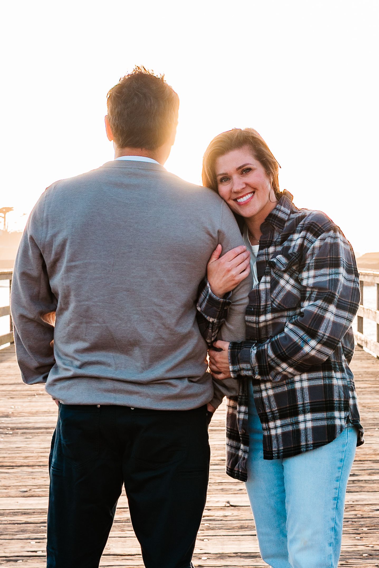 Smiling woman in plaid shirt embraces man from behind on a sun-drenched wooden pier during golden hour.