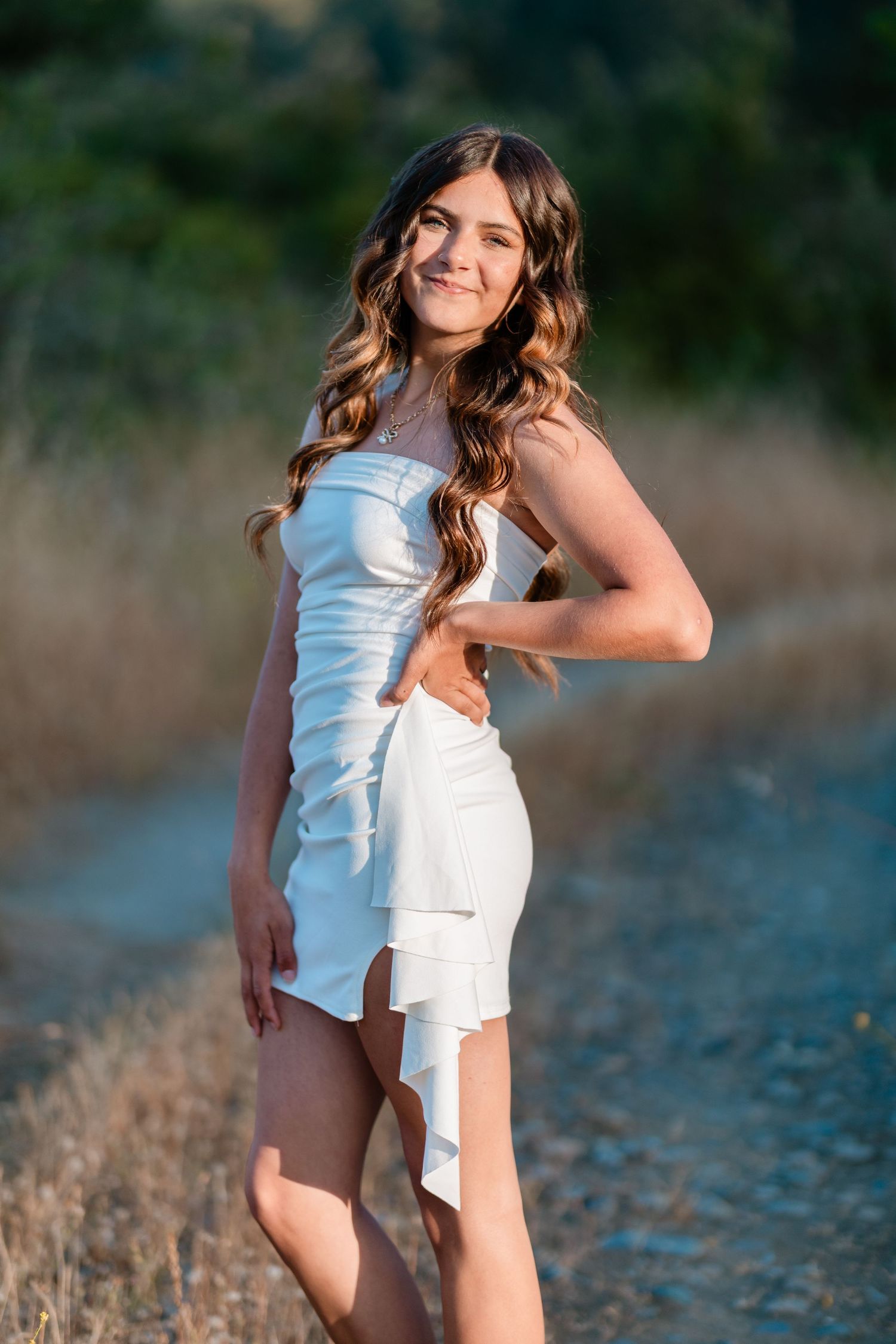 Young woman in white mini dress posing on a sunny dirt path surrounded by golden grass and green trees.