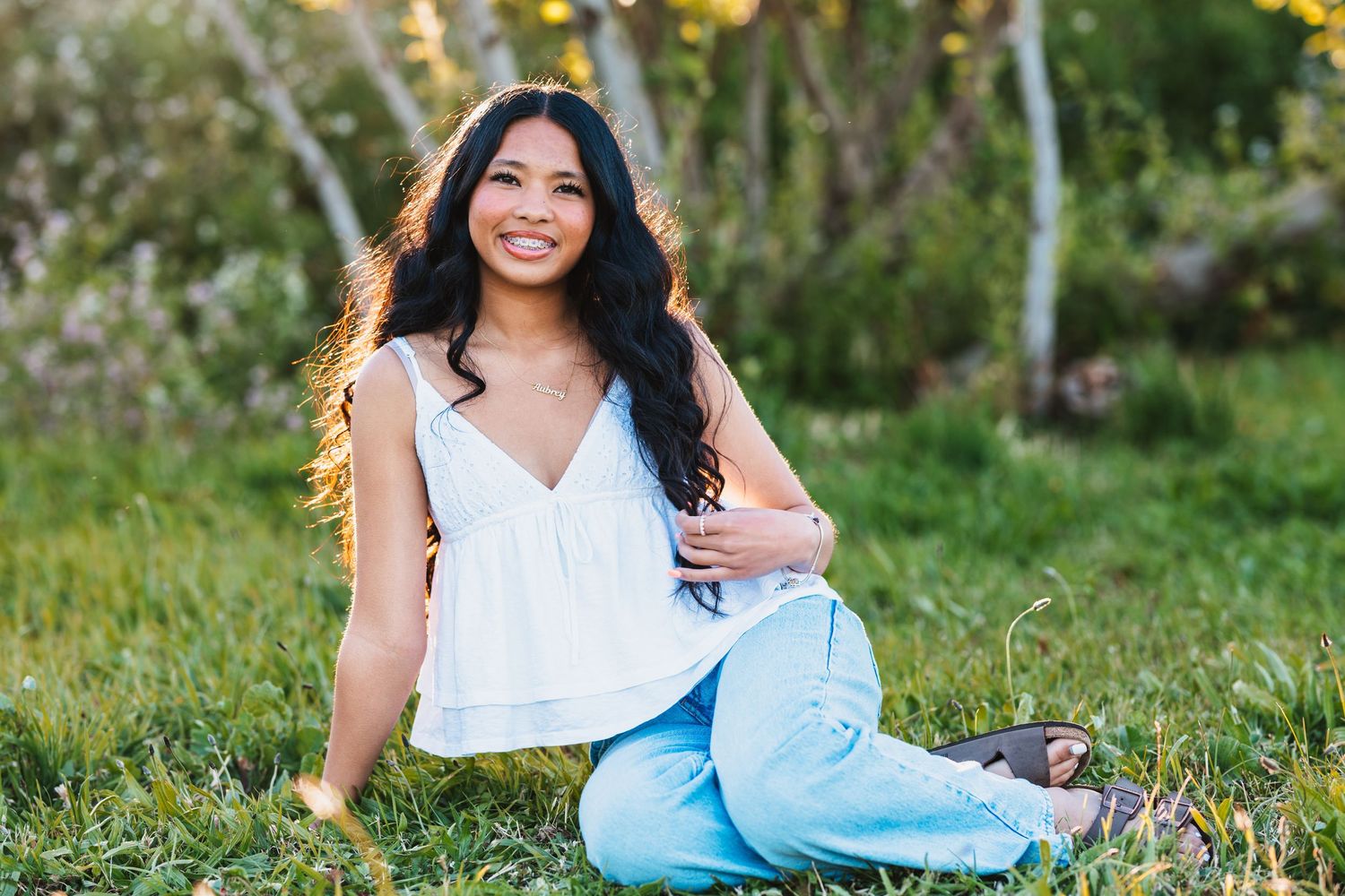 Young woman with long dark hair smiling, sitting on green grass in white top and light blue jeans outdoors.
