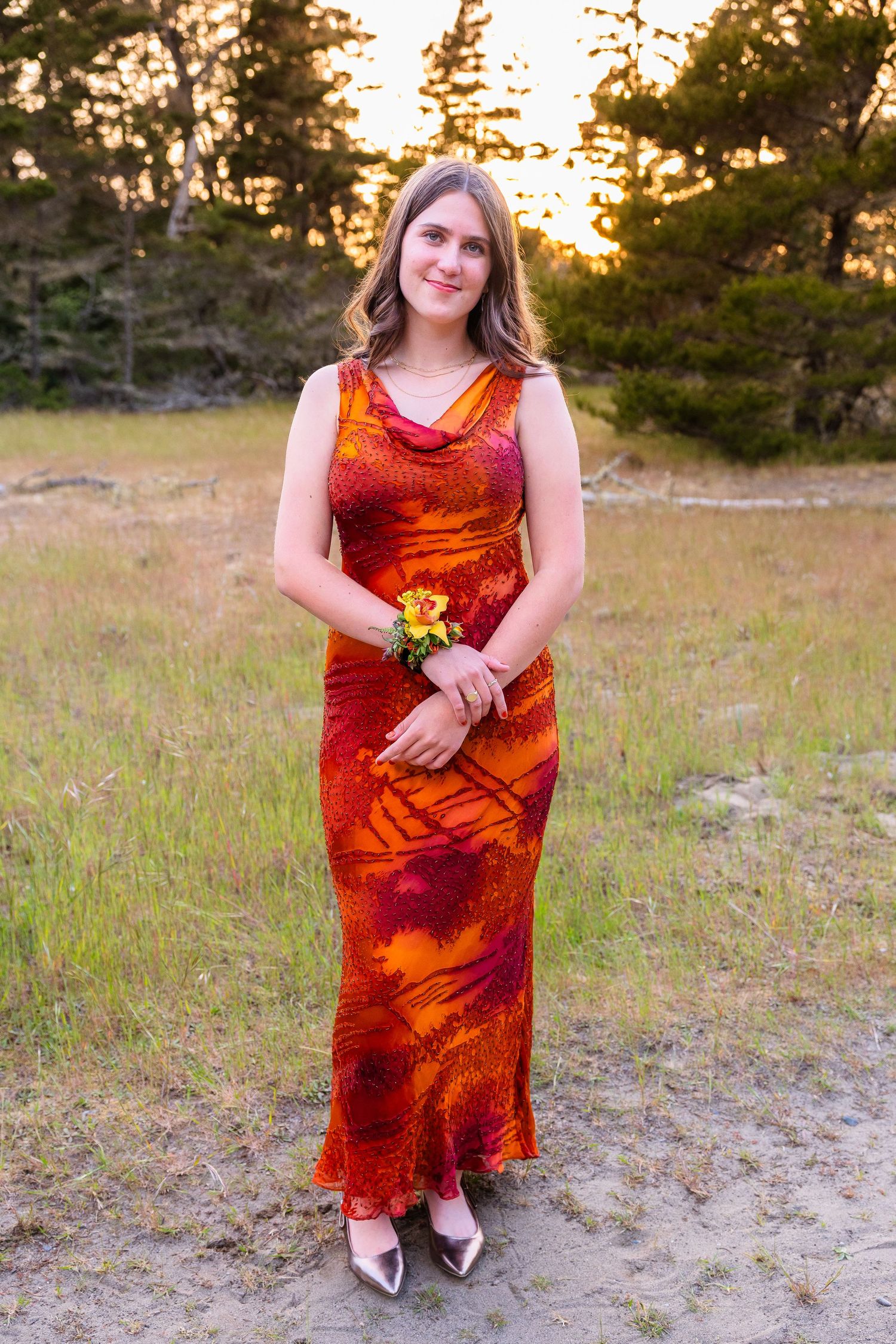 Young woman in red orange floral gown holding corsage poses outdoors at sunset near pine trees.