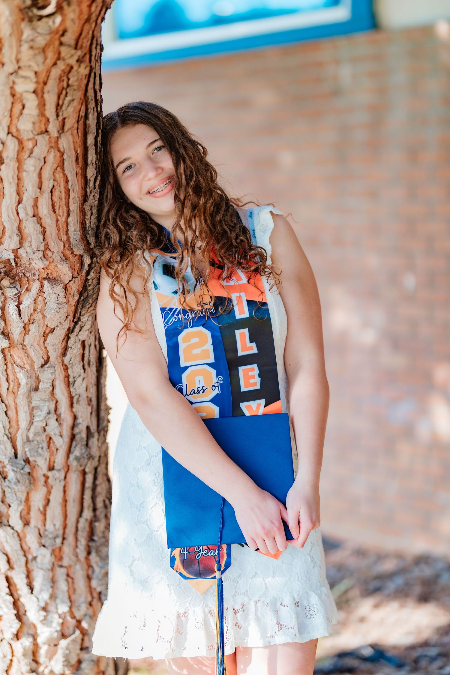 Teenage girl in white dress holding graduation stole leans against tree trunk outdoors smiling.