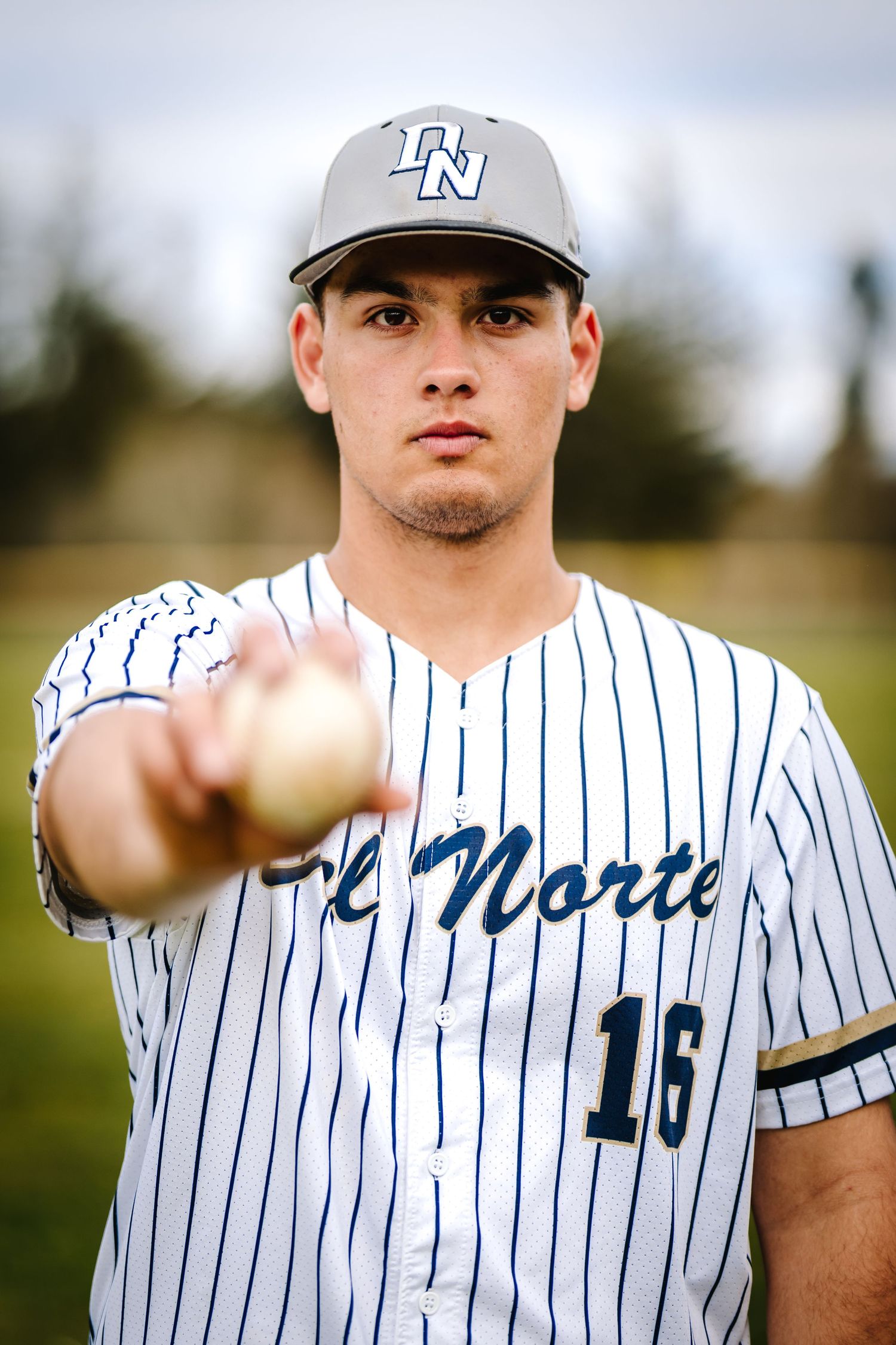 Del Norte baseball player #16 in pinstripe jersey holding a baseball toward the camera.