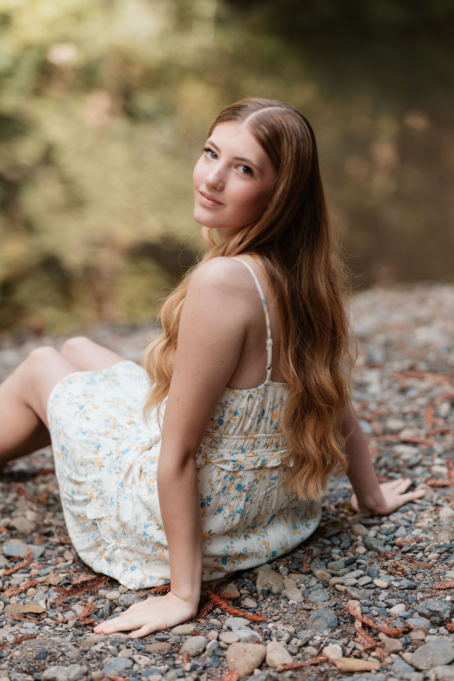 Young woman in floral sundress sitting on rocky riverbank, looking over shoulder with long wavy hair.