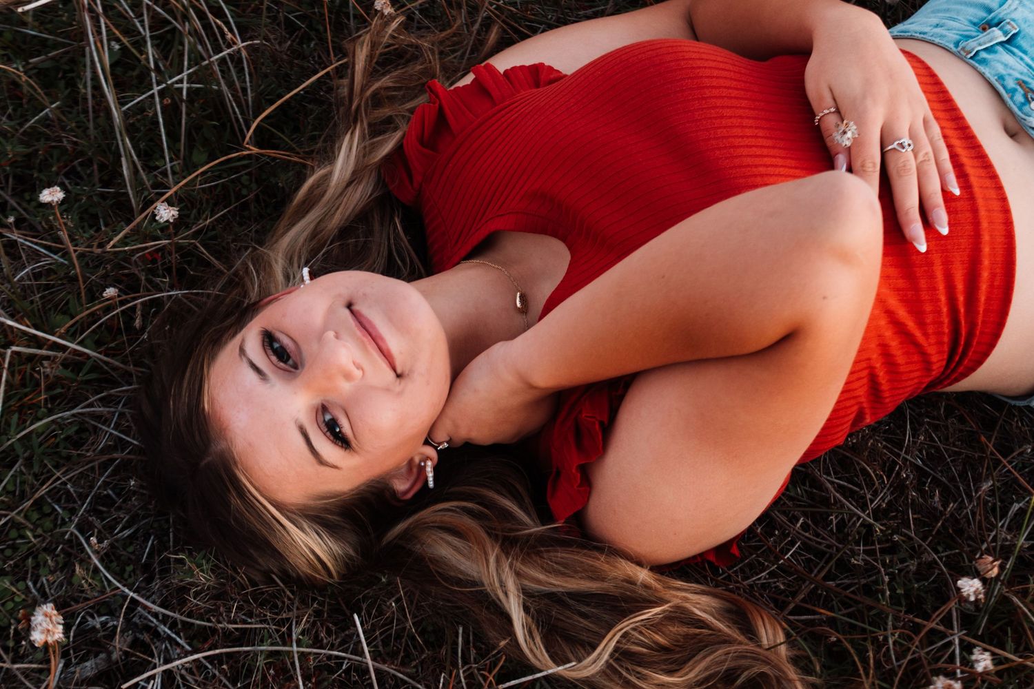 Teen girl in red top lying in grass smiling up at camera during outdoor senior photo session.
